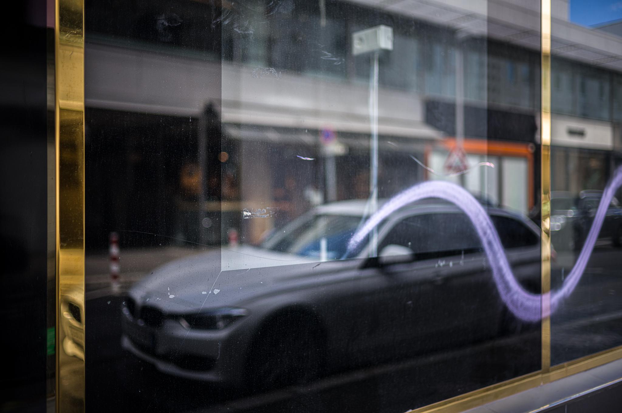Empty (except for two golden thin poles) shopwindow with a graffito-wave, mirroring opposite buildings and a parking BMW.