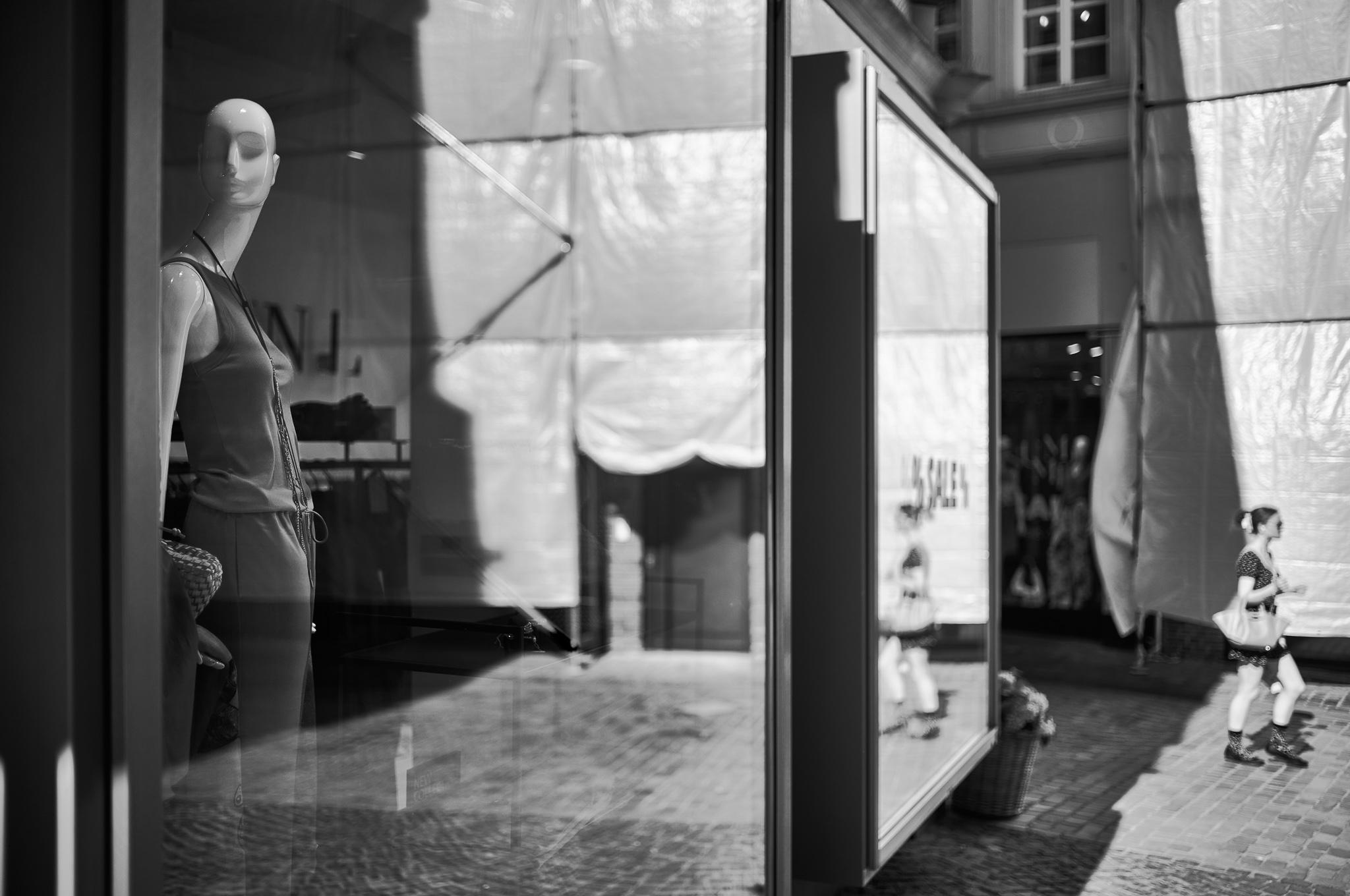 B/W photo of a shopwindow partly reflecting opposite building which is covered by scaffolding. Near the left edge there is mannequin in the shopwindow looking to the left , at the far right a blurry person is walking towards the right