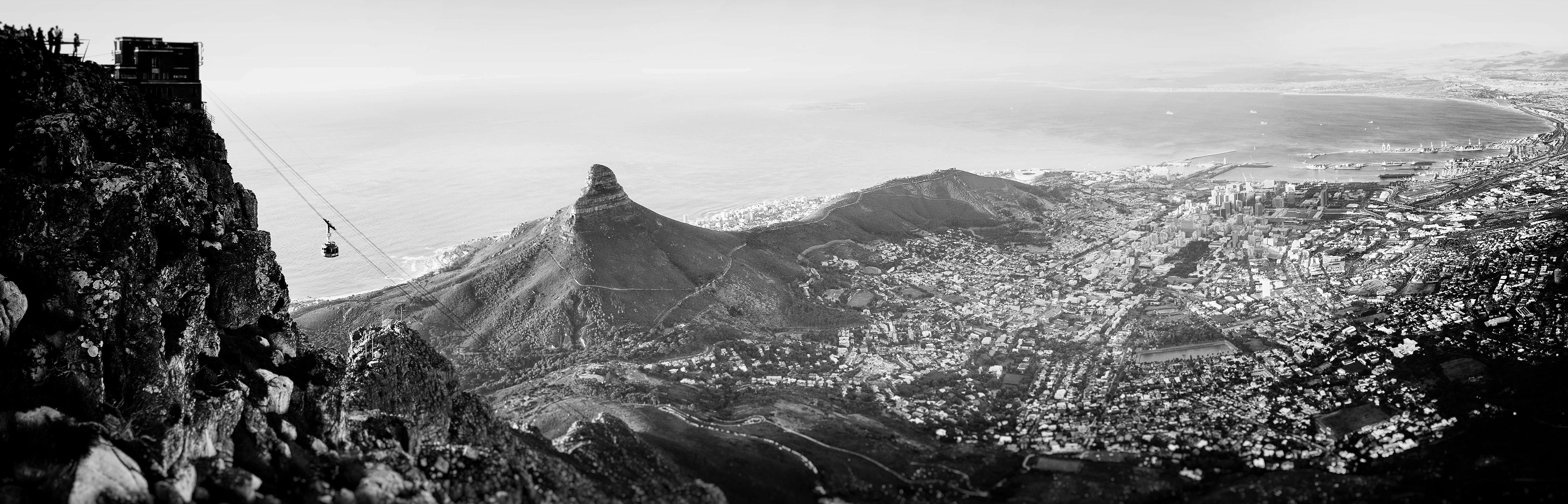 Panoramic black-and-white view from the top of Table Mountain in Cape Town, showing the cable car descending toward the city, Lion’s Head peak, and the Atlantic Ocean coastline