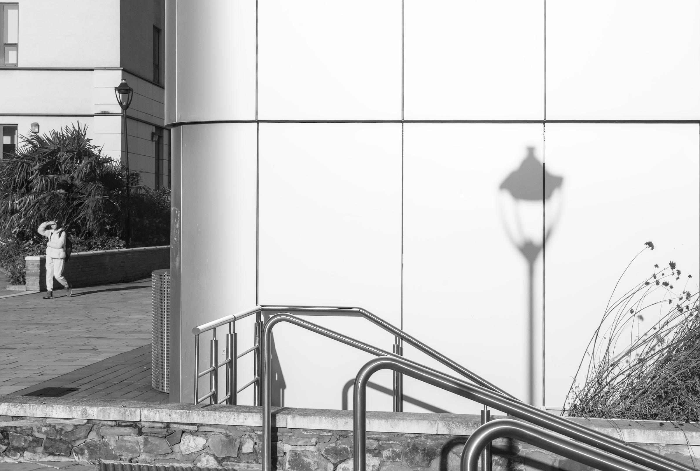 A black and white photgraph taken at Bristol University, St Michaels Hill, Bristol UK.
The image is of, a modern metallic silver sturcture which is reflecting strong sunlight. There is a shadow of an old gas style lamp falling on the building at the right of centre frame.

The forground is of a low stone built wall with smooth capping stones and also bright stainless steel handrails belonging to steps that are out of the frame.

To the far left, of the frame is a young woman shielding her eye from the sunlight as she walk forward.