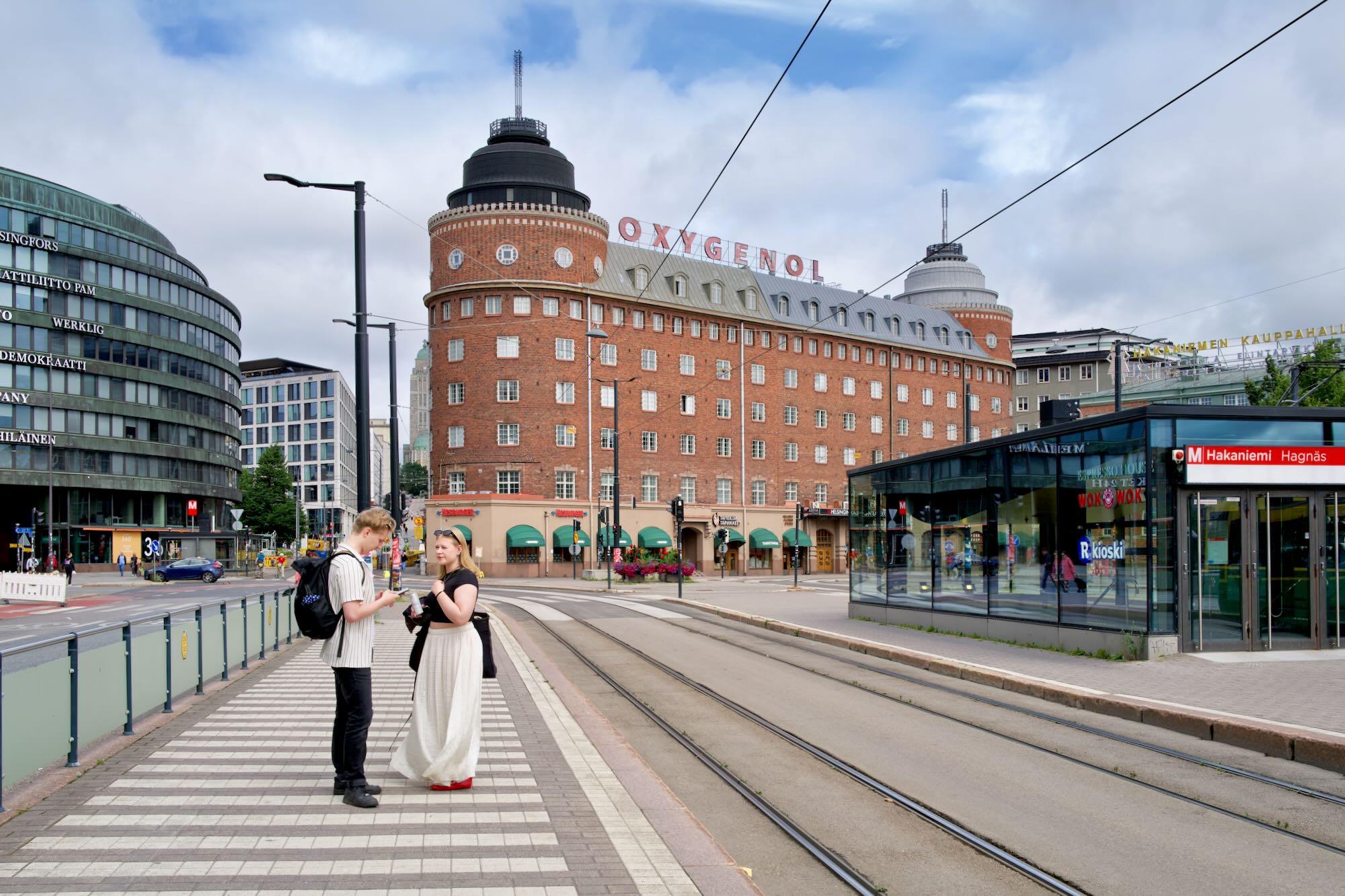 1. A broad city street in Helsinki is seen beneath an overcast sky, with tram tracks curving through the foreground.
2. A large red-brick building with two distinctive cylindrical towers dominates the center, topped by the word “OXYGENOL.”
3. Modern glass-and-steel buildings surround it, including a glass-covered tram or metro entrance marked “Hakaniemi.”
4. Two pedestrians stand on the tiled sidewalk in the foreground, while cars, street signs, and traffic lights occupy the surrounding intersection.
5. Overhead tram wires stretch across the sky, emphasizing the urban setting and public-transport character of the scene.