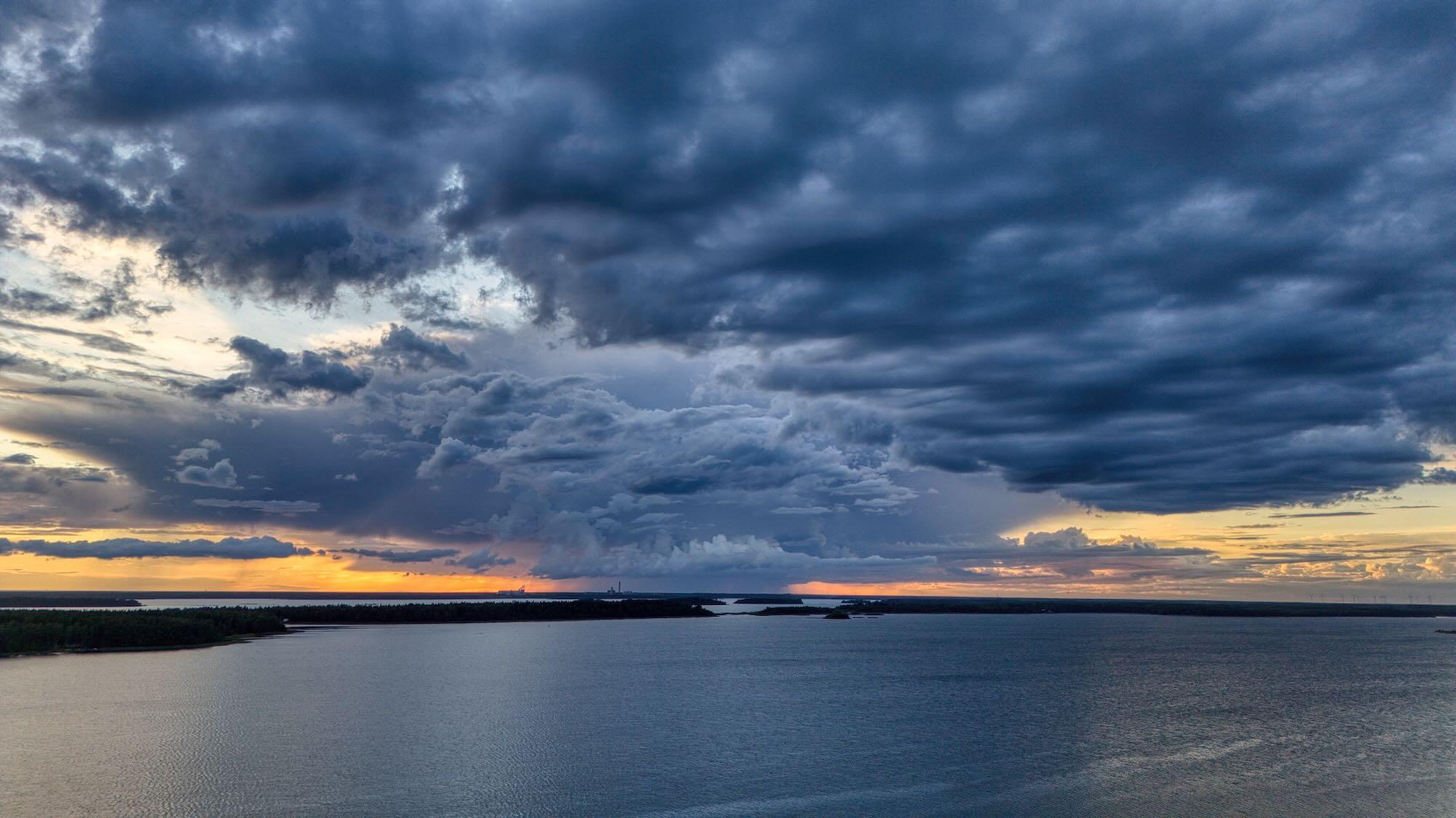 A vast body of calm water stretches toward a flat horizon, dotted with low, dark islands.
The sky is filled with dramatic, layered storm clouds in deep blue and gray.
Near the horizon, a band of warm golden and orange light suggests sunset.
The water reflects the fading light, creating a peaceful contrast with the dark clouds.
The scene has a vast, atmospheric feeling, with land, sea, and sky dominating the view.