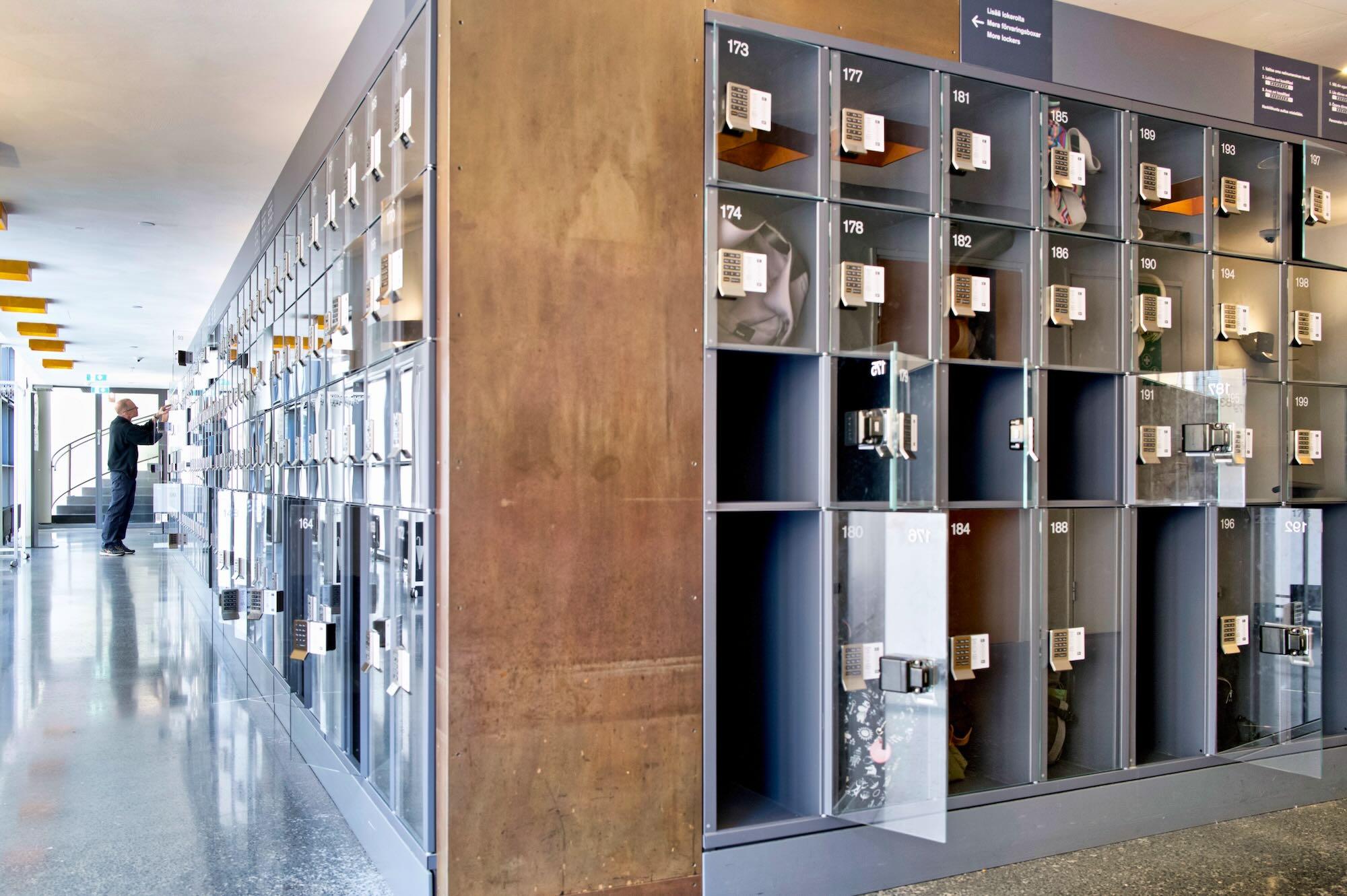 Visual description

A long, modern corridor is lined with hundreds of small numbered storage lockers arranged in neat rows.
Each locker has a dark interior and a keypad-controlled lock, with some compartments open and containing personal belongings.
The perspective makes the lockers appear to extend far into the distance along the left side of the corridor.
A person stands near the far end, reaching toward one of the lockers, while glass doors and a staircase are visible behind them.
The polished floor reflects the lockers and bright overhead lighting, creating a clean, orderly and slightly futuristic atmosphere.