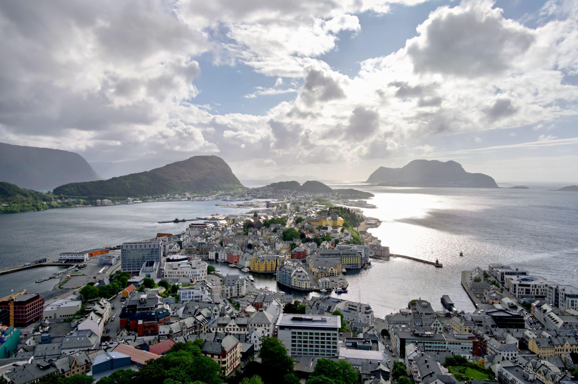 A high panoramic view looks down over a dense coastal city surrounded by water and dramatic mountains.
Colorful buildings and gray rooftops cluster around a harbor filled with small boats and waterfront docks.
A narrow peninsula extends into the water, with steep green hills rising behind the city.
Bright sunlight reflects intensely off the sea beneath a sky filled with large, luminous clouds.
The combination of compact urban life, mountains, islands, and open water creates a striking sense of scale and place.