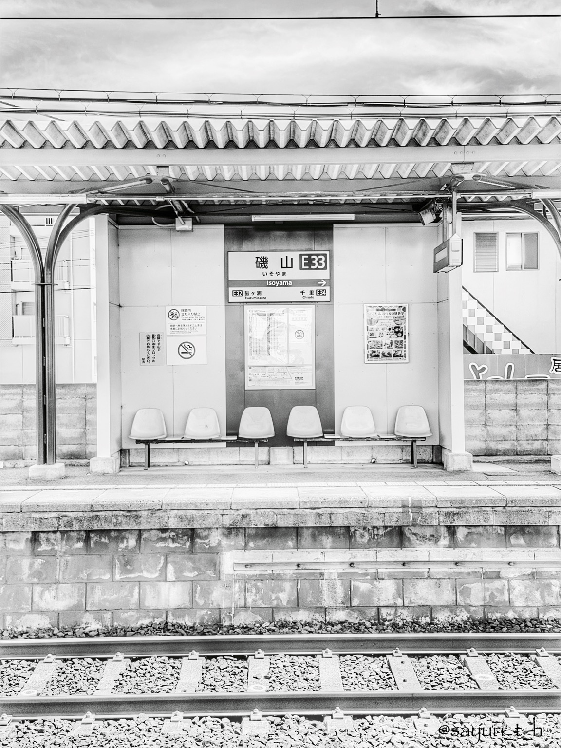 Black-and-white photo of the quiet platform at Isoyama Station in Japan, with empty seats beneath a covered shelter and railway tracks in the foreground.