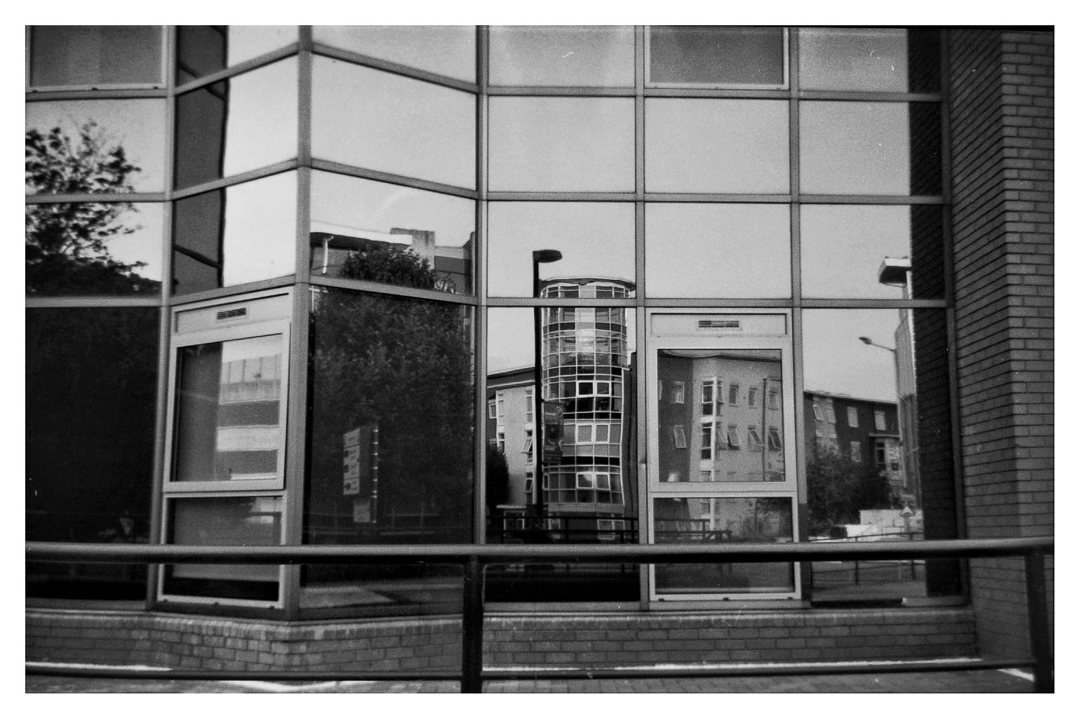 A (slightly less) grainy black and white image of some apartment buildings reflected in the windows of an office block.