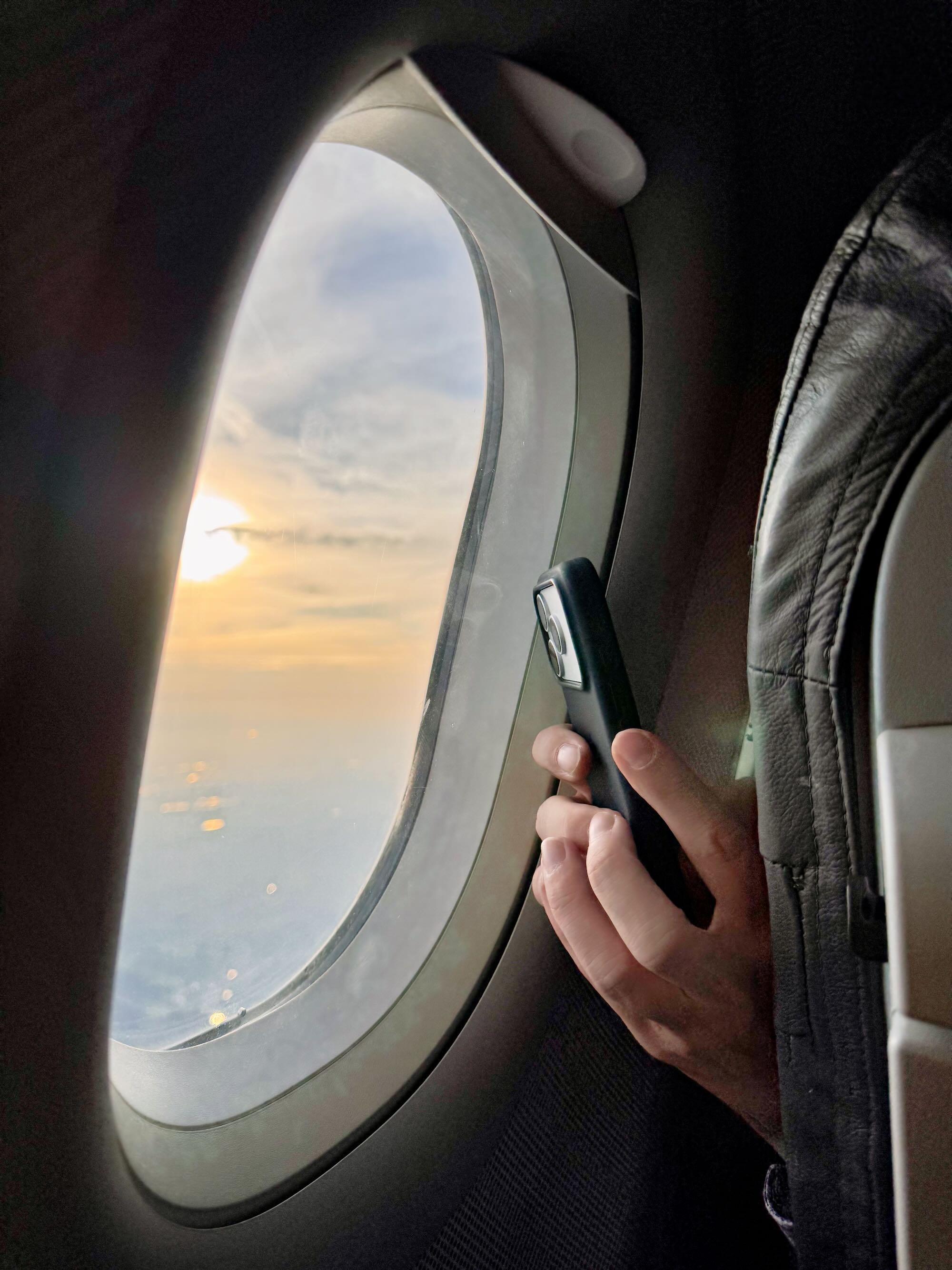 A close-up view from inside an airplane shows an oval window with a hazy sunset glowing outside.
A passenger’s hand holds a smartphone vertically beside the window, apparently photographing or filming the view.
The airplane seat and dark leather headrest frame the right side of the image.
Soft golden light from the setting sun contrasts with the dim interior of the aircraft.
The image conveys a quiet moment of travel, observation, and anticipation.