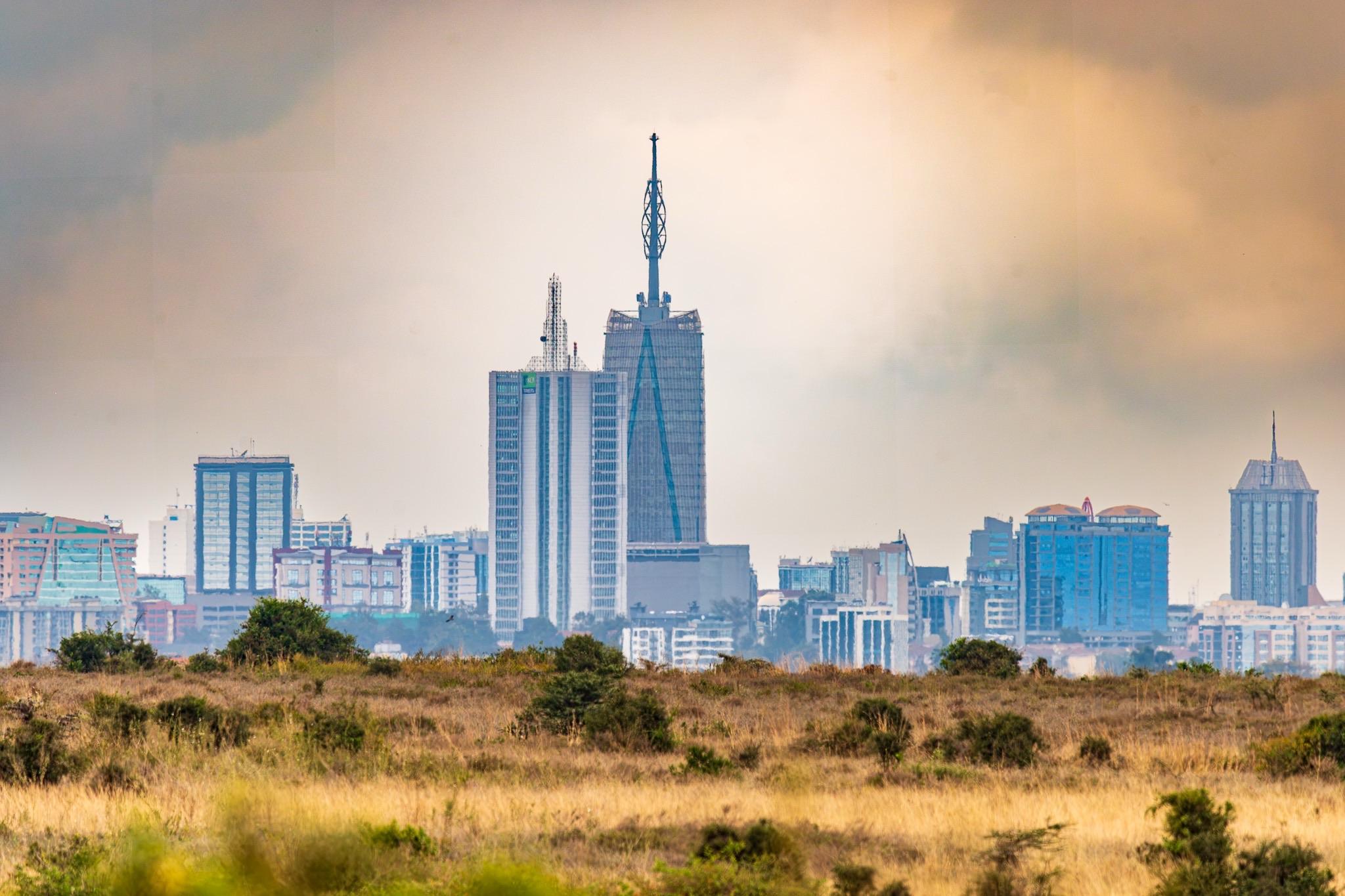 A skyline view of a city featuring several tall buildings, with one prominent tower topped by a unique antenna. The foreground consists of grassy land, and the sky is overcast.
