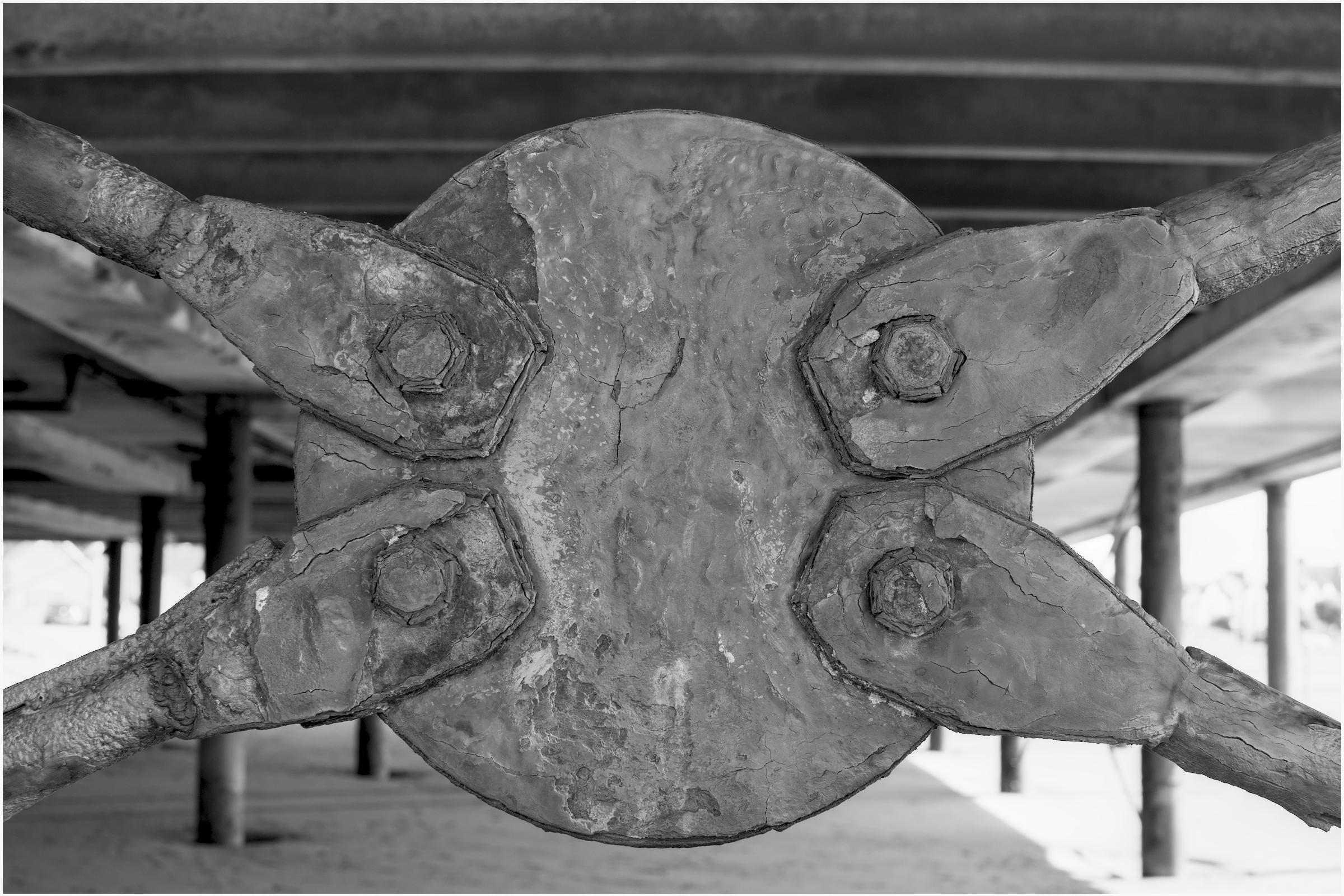 A black and white photograph of an iron brace, a disc with drilled holes to which are bolted four stays or tie bars)' under a small pier in Weston Bay, WsM, Bristol, UK.

Time and tide are graually having a corrosive effect on these parts. Though one images the engineer anticipated allowed for this degradation.