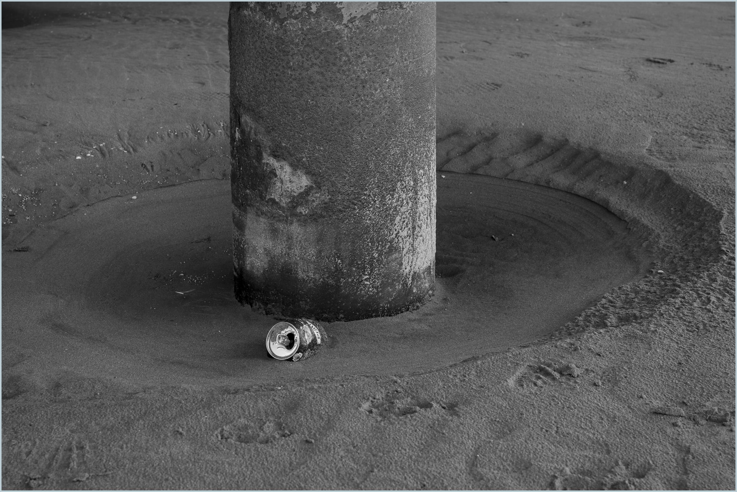 A black and white photograph of an iron piling, in a crater of sand, under a small pier in Weston Bay, WsM, Bristol, UK. In the crate is also a discarded and dented beer can, which is still shiny new. Whereas the pillar, being the subject the element from the Bristol Channel, has all the signs of coatal corrosion