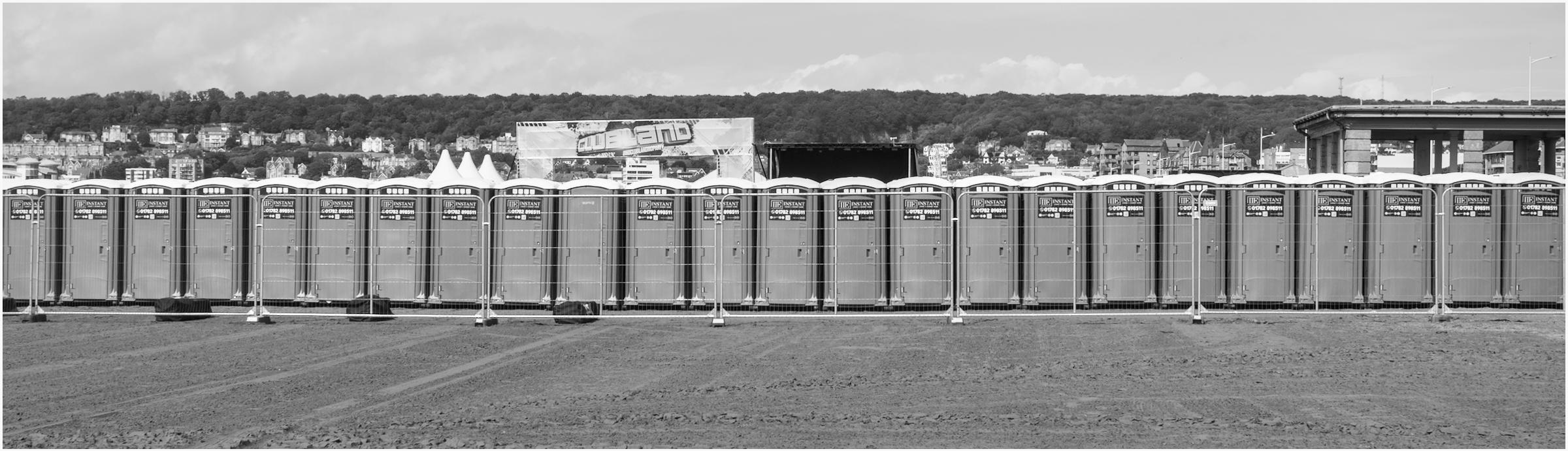 A black and white letter-box shaped photograph of twenty four Instant Loos taken on Weston Beach, Weston Bay, Bristol, UK. The tenth loo from the left is the odd one out - it has no self advertising sticker.