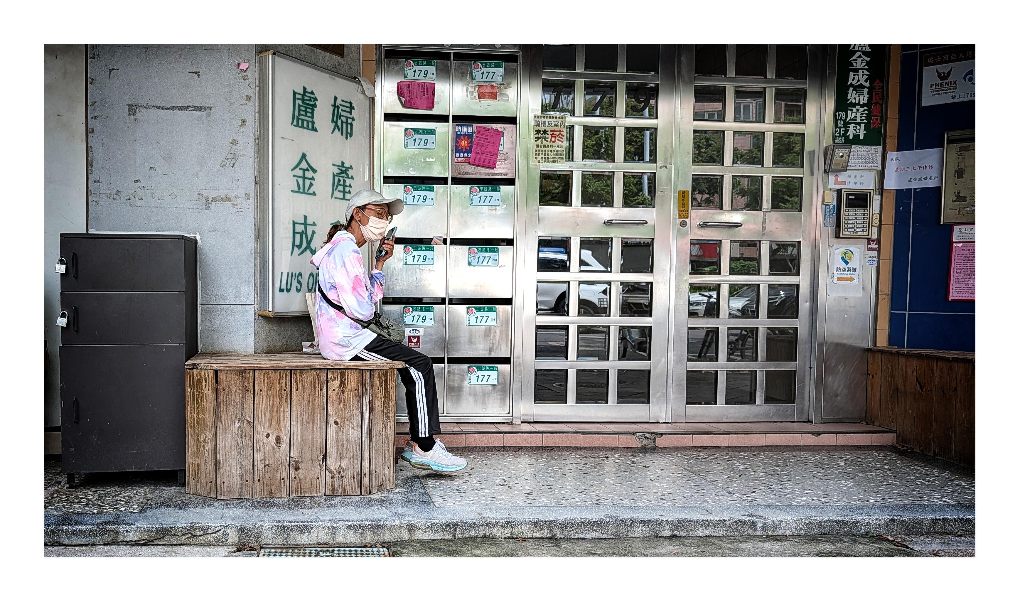A masked woman sits on a wooden box outside an older building in Taipei, holding a smartphone close to her face. Rows of metal mailboxes and reflective doors fill the background, with Chinese signs for an obstetrics and gynecology clinic adding to the everyday urban setting.