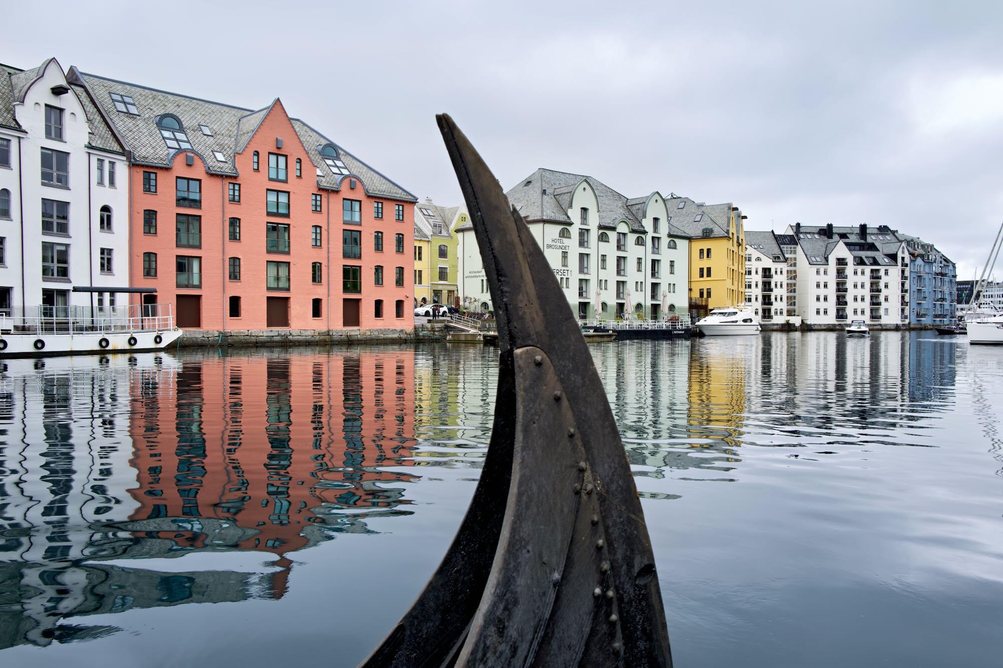A replica Viking ship’s dark wooden bow rises prominently from the water in the foreground of Ålesund Bay.
Behind it, colorful waterfront buildings with steep, slate-covered roofs line the calm harbor.
White, salmon, yellow, and pale-blue façades create a vivid contrast with the gray, overcast sky.
The buildings and boats are reflected in the gently rippling water, creating distorted vertical patterns.
The composition juxtaposes maritime heritage with Ålesund’s distinctive historic waterfront architecture.