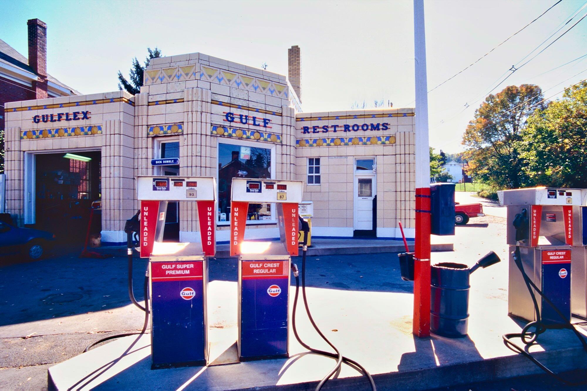 A vintage Gulf gas station is photographed from the forecourt in bright sunlight. Three old-fashioned fuel pumps stand prominently in the foreground, painted blue and red with white panels and hoses. The pumps are labeled “Gulf Super Premium,” “Gulf Crest Regular,” and “Gulf Crest Regular.” Behind them is a low, cream-colored Art Deco–style service station building decorated with geometric blue, yellow, and red patterns. Large red-and-blue lettering reads “GULF” and “REST ROOMS.” A red-and-blue Gulf signpost and a blue metal barrel stand to the right. Trees, overhead utility wires, and residential buildings are visible in the background. The strong sunlight creates long, dark shadows across the pavement, giving the scene a distinctly nostalgic, mid-20th-century American roadside atmosphere.