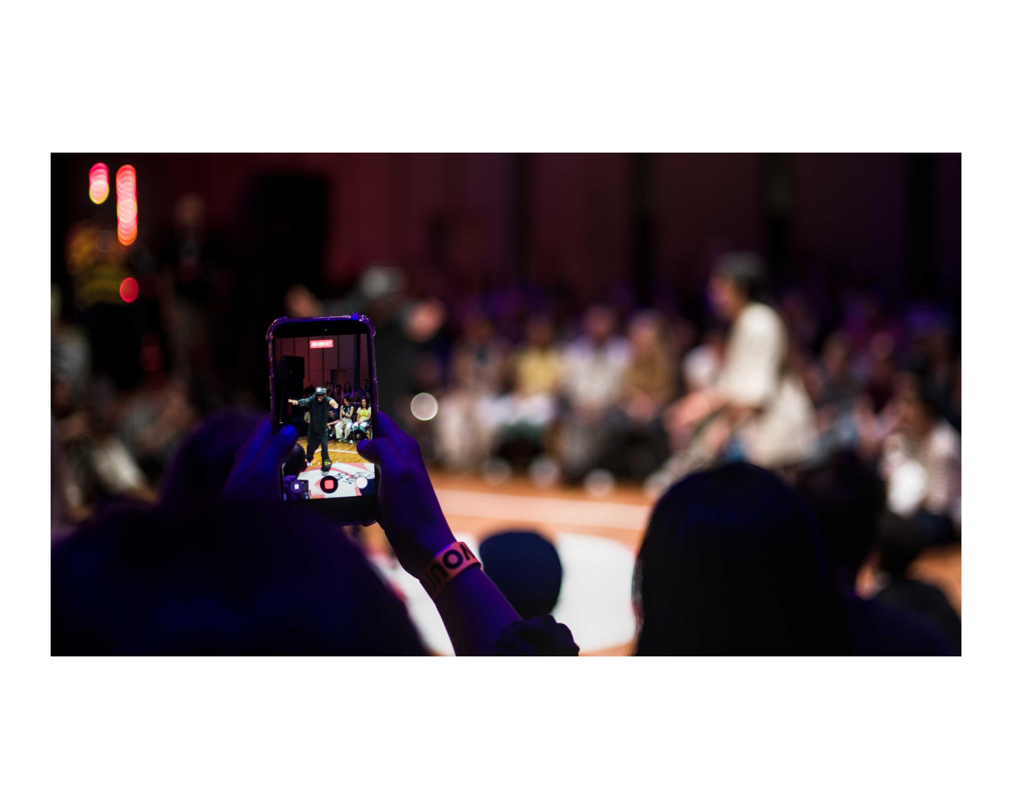 A dance battle in background, in the light but also out of focus. A dancer is seen in focus from the screen of a phone, held up to record the dancing. Heads of people, dark stand in foreground framing the picture. More audience is behind the dancers, opposite from where the picture is taken.