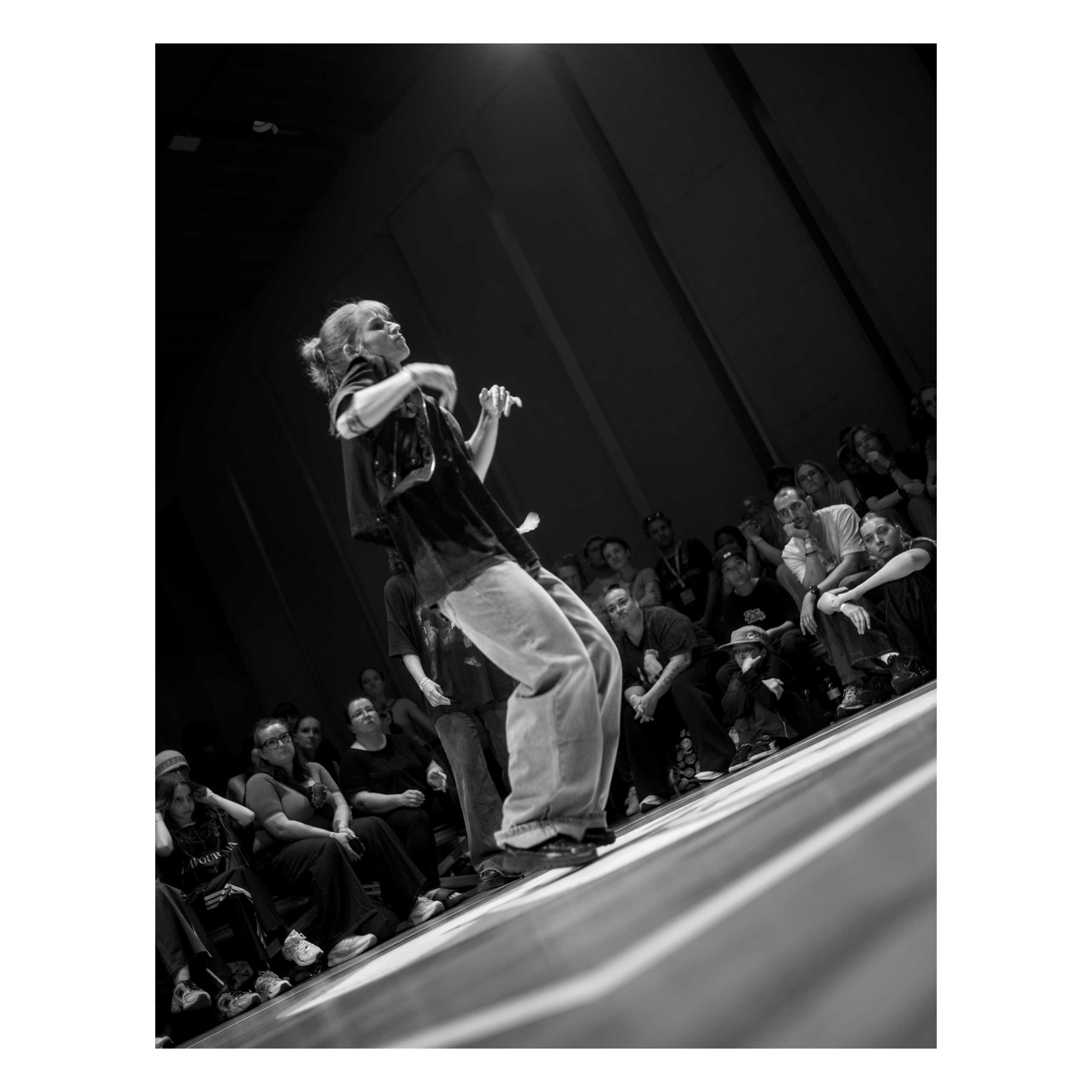 A black and white picture of a dancer, standing on the point of her shoes, knee bents, arms sharp out and perpendicular to the floor. The picture is taken with a tilt angle to the left, and the light shine stark from above. The dancer is wearing light jeans and a dark t-shirt and gilet. The public is looking in background.