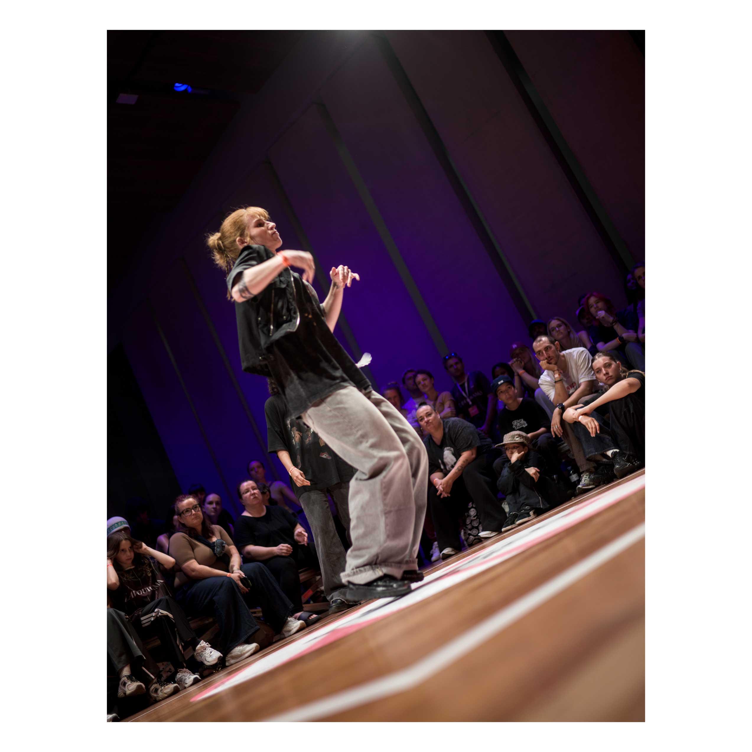 A color picture of a dancer, standing on the point of her shoes, knee bents, arms sharp out and perpendicular to the floor. The picture is taken with a tilt angle to the left, and the light shine stark from above. The dancer is wearing light jeans and a dark t-shirt and gilet. The public is looking in background.