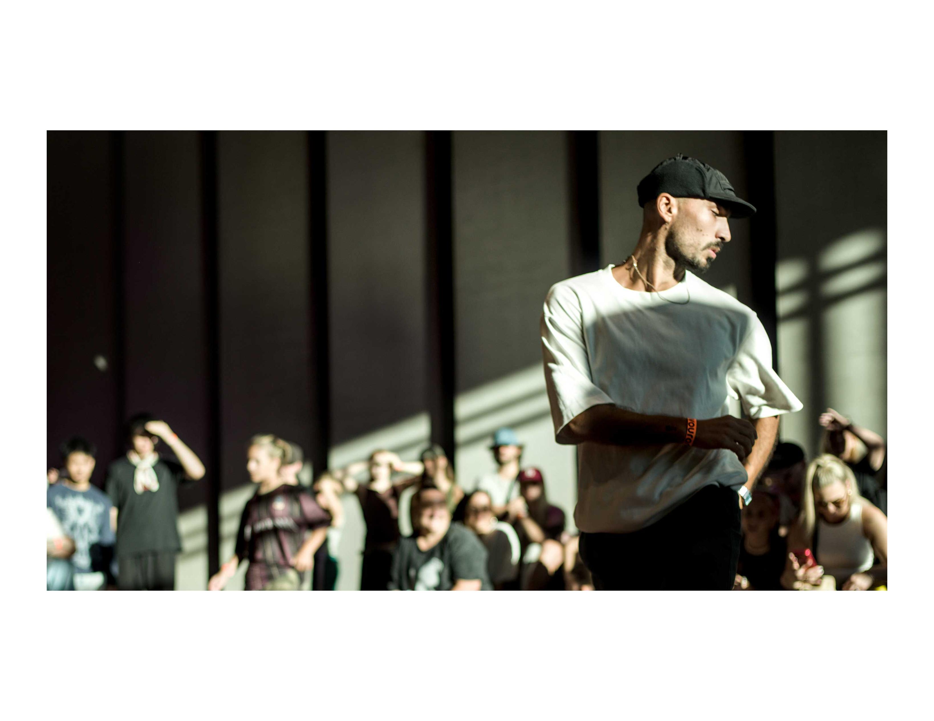 color photo with a male dancer on the right and the public out of focus in background. The sun's light is coming sharp from the left, angled toward the left bottom angle. The light plays on the dancer, which is turning to the right, the chest is still facing the camera but the head is looking to the side and the arms are also moving.