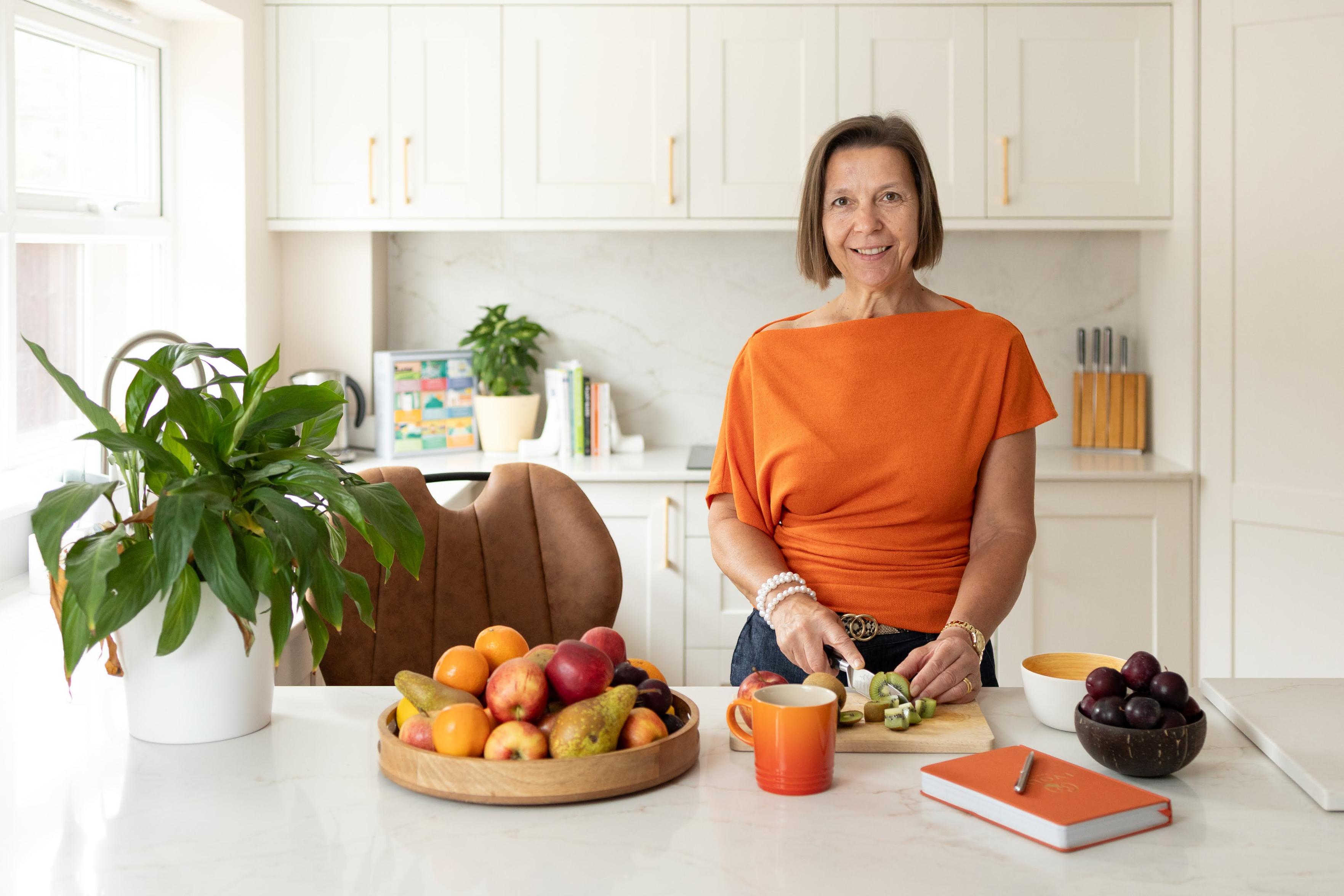 Environmental portrait of a woman cutting fruit in a kitchen. It's a bright white kitchen. The woman is wearing an orange top, with orange also picked out on the worksurface with a book, cup and fruit. The woman is smiling and looking directly at the camera.