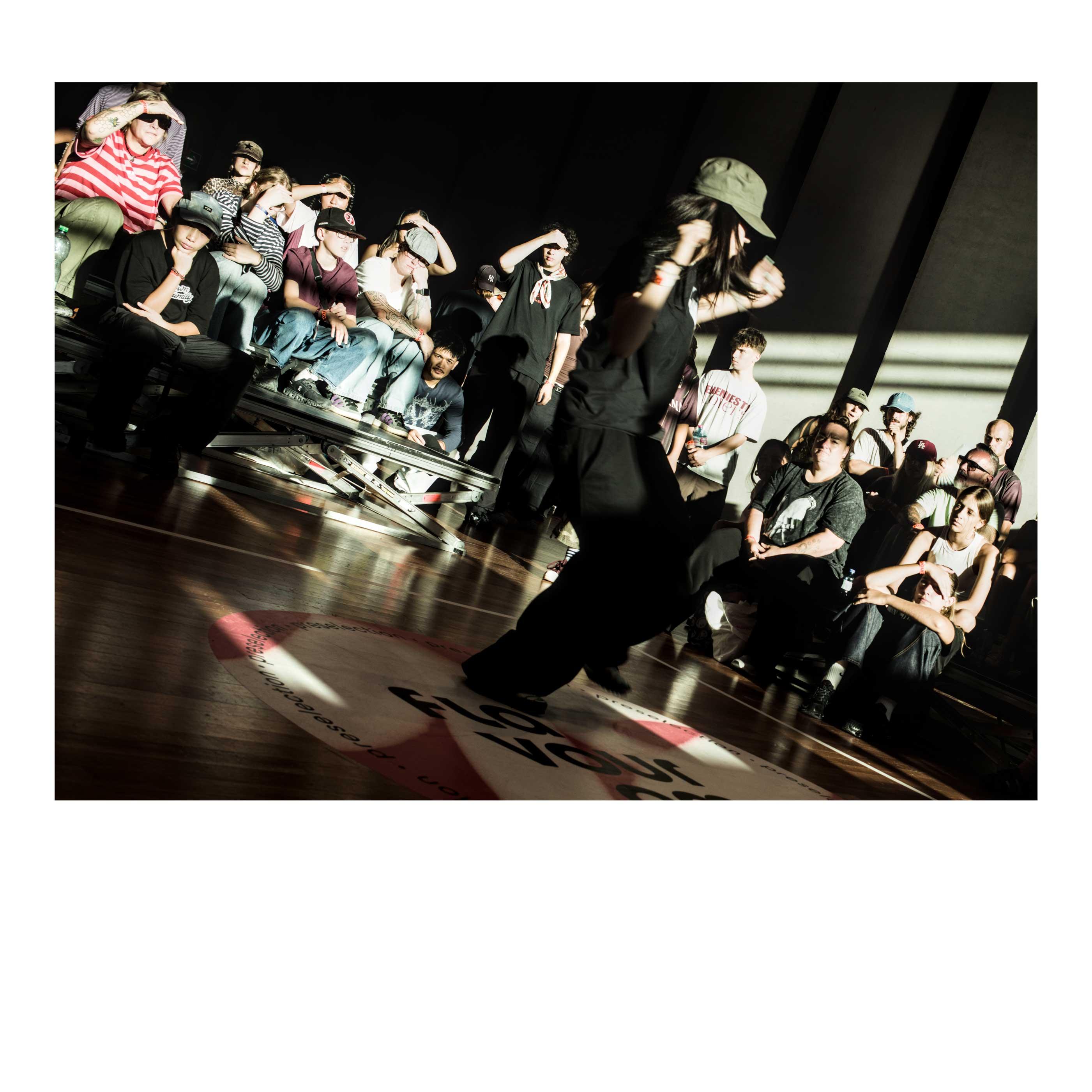 A dancer, in black clothes and military green hat, dark long hair, slightly bending her knees as her arms are moving. She is angled toward right of the picture. The public behind is looking with all their eyes locked on the dancer, some shielding their eyes from the sun coming in sharp from the right, cutting lines of light and dark in the picture. On the floor the Flavourama logo.