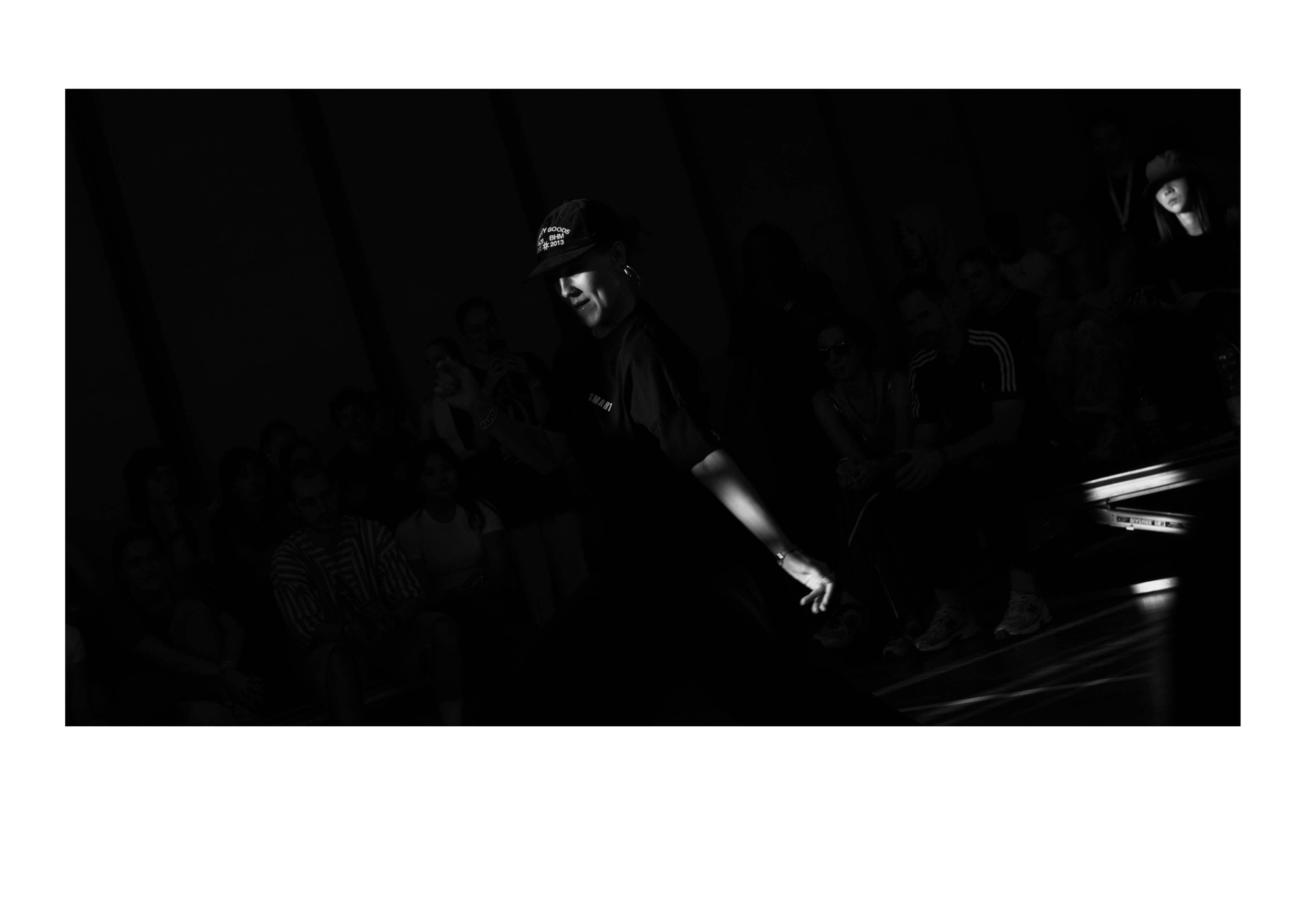 black and white picture of a dancer. However, the black is the dominant color, with only the face with a hat and the left arm of the dancer in the light, together with a face far in the back. The hat has white writings.