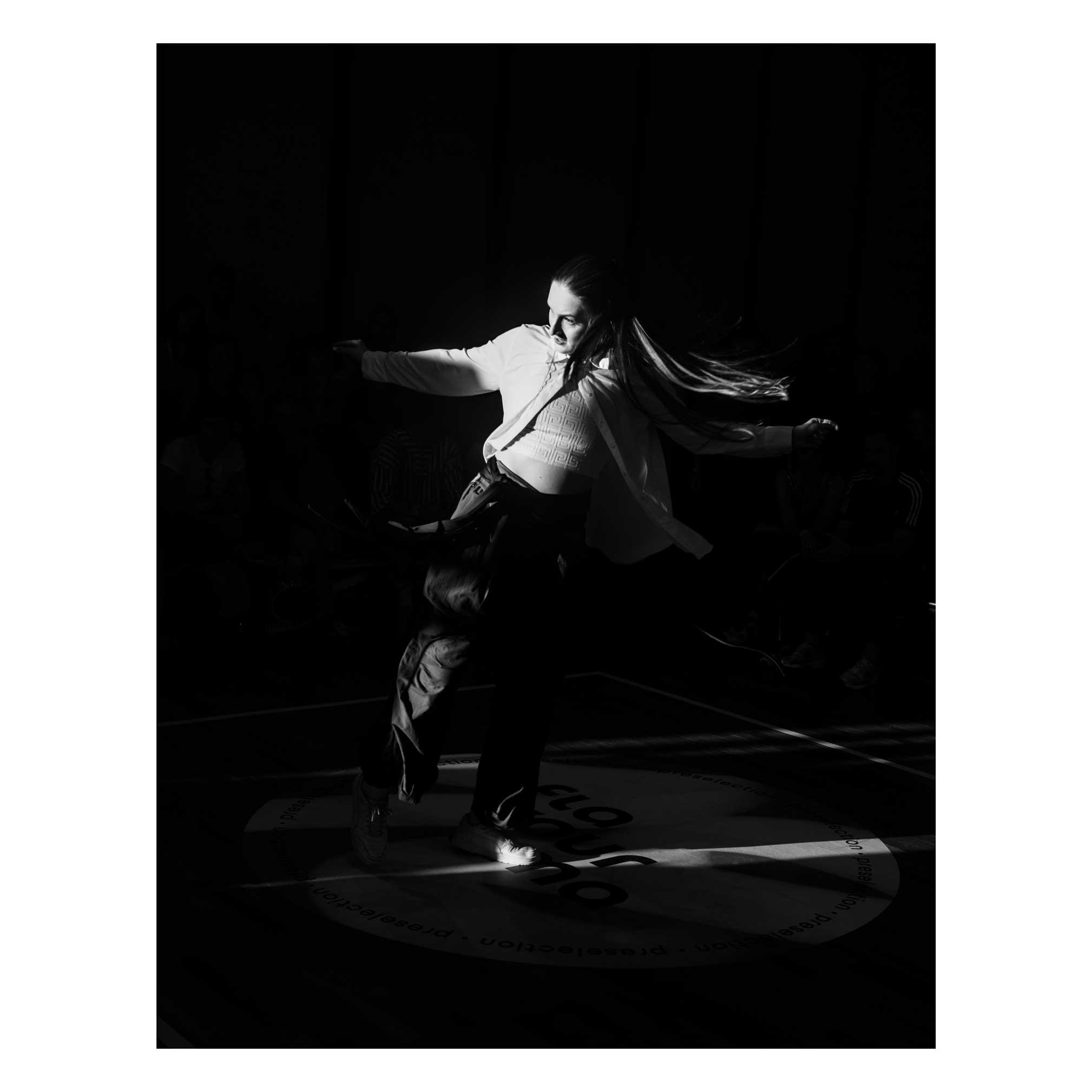 A black and white picture of a house dancer stepping into a slice of light coming in from the left. Her hair are flowing behind her, left foot forward, right foot raised, arms spread. The light only shine on her upper body, lightening the emotions on her face.