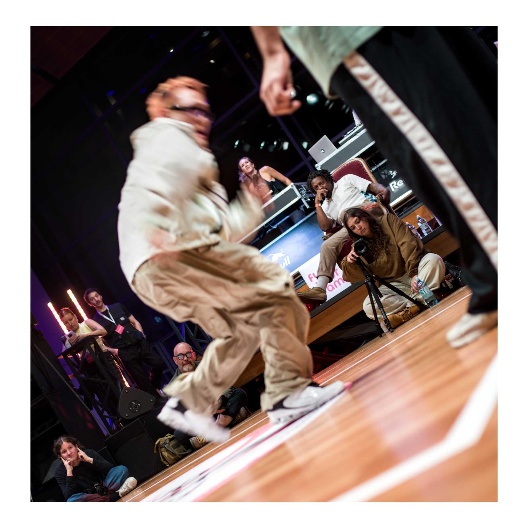 A color picture of a house dancer. The dancer is blurred, in fast movement. The background, however, is sharp and in focus, with all the eyes set on the dancer. Photographer, dance judges, videographers, even the DJ is looking at the dancer. The angle is tilted to the left. On the right, the out of focus black pants with a thick while line of another dancer waiting his turn.