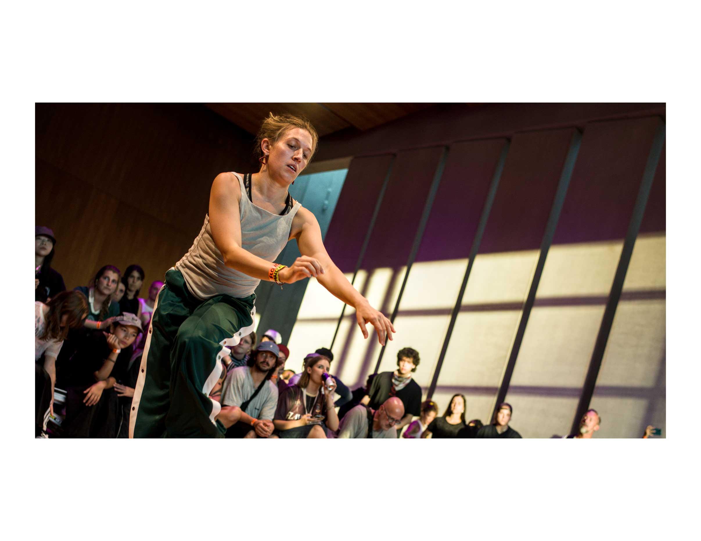 A photograph in color of a house dancer. The dancer, has her left hand forward, as if to pick something with the tip of her fingers. She is wearing dark green pants with a white line and a grey tank top. Her eyes are on the invisible object she is picking. The onlookers, behind, are slightly out of focus. The colors are farm, with the shine casting straight, sharp lines on the far wall.
