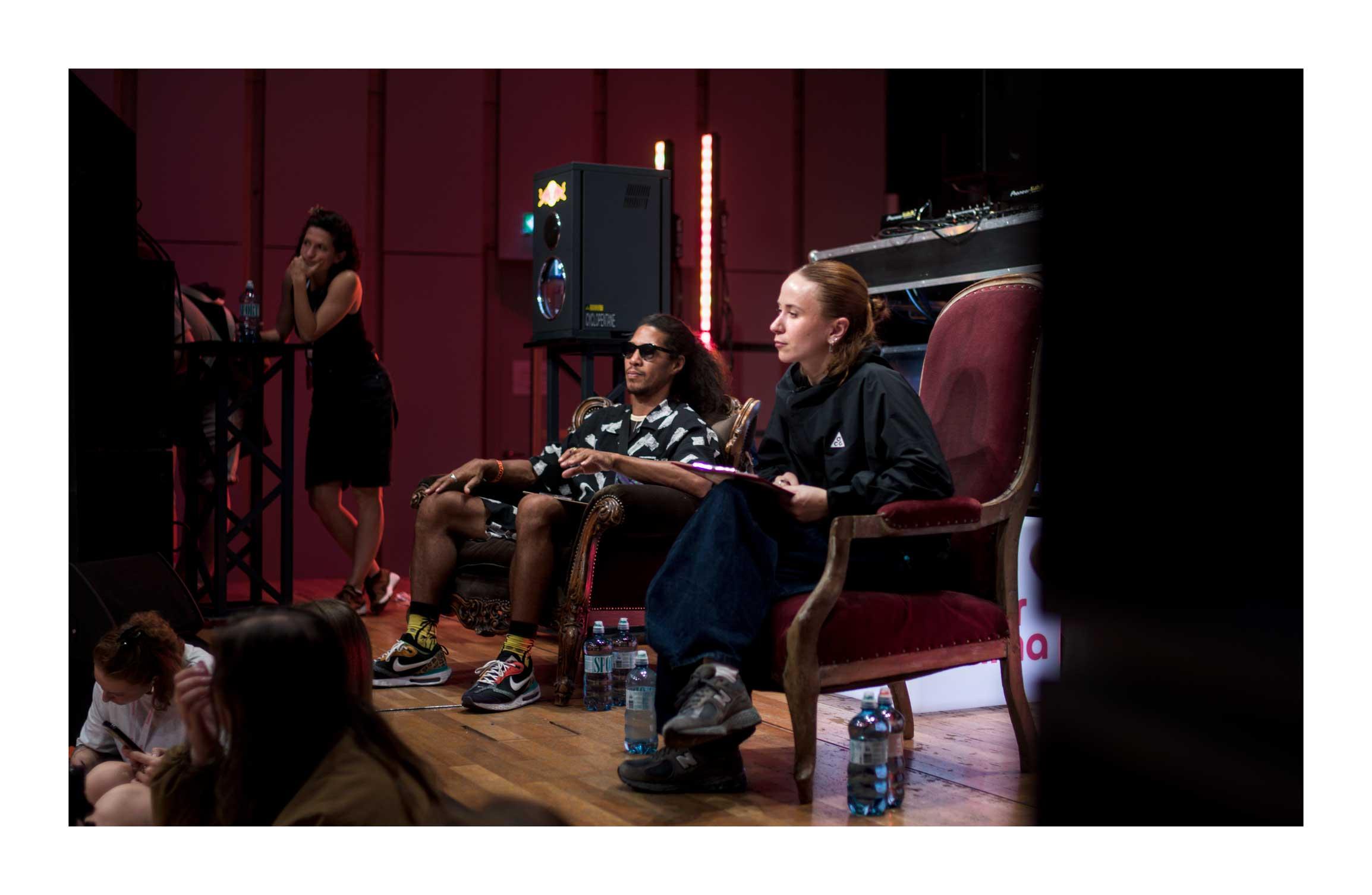 A color photography of two of the judges of a dance battle. The one closest to the camera, with dark pants and legs crossed, is leaning forward looking attentively at the battle out of the picture. The other judge, sits much more relaxed with shorts and sunglasses. At the far end, one of the staff in black leans on a tall bar table with her elbows as she too watches the battle.