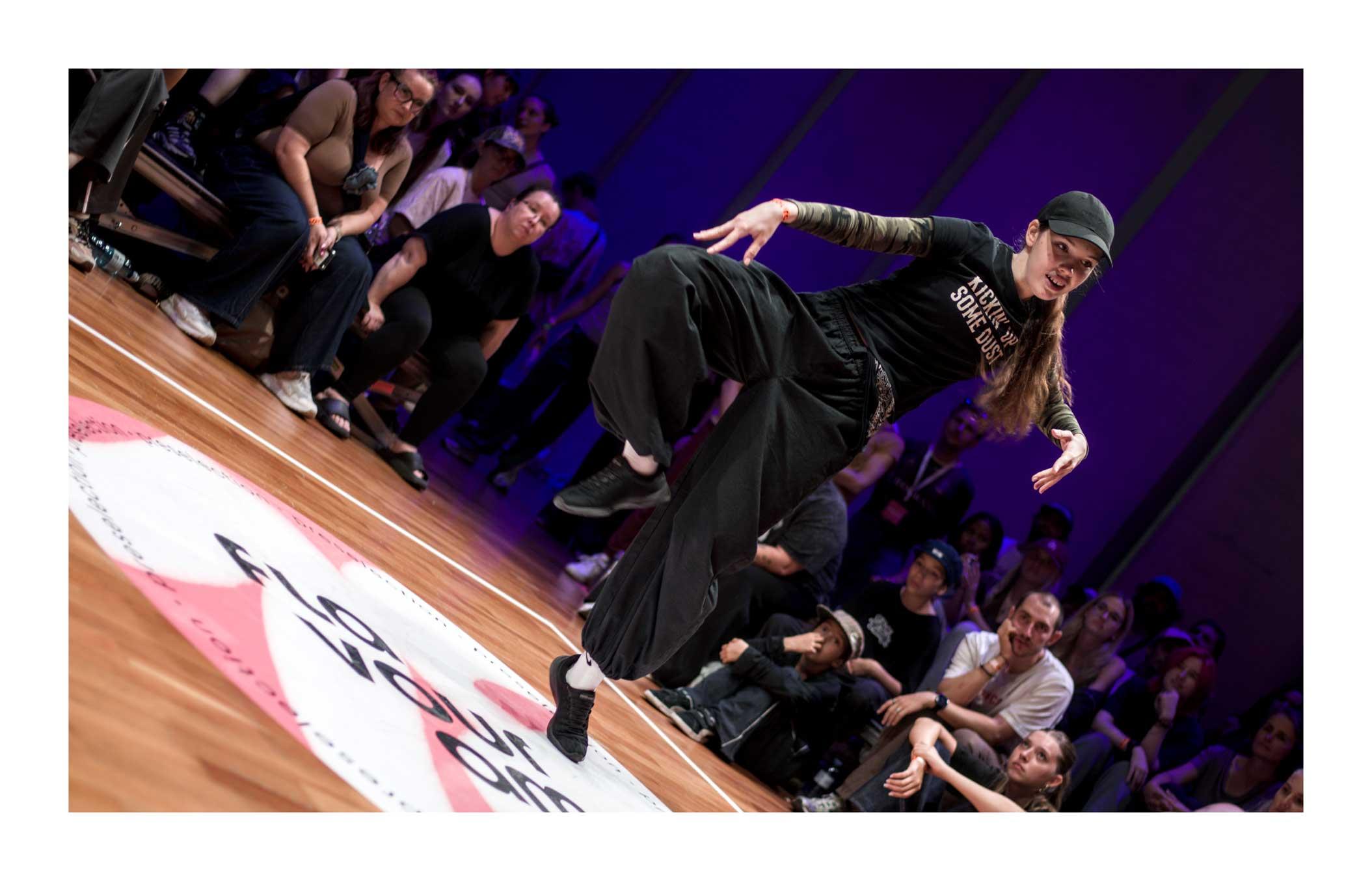 A color photograph of a dancer during a dance battle. She is standing on the point of her left foot, right raised and bent at the knee. The arms are spread with hands pointing down. It is akin a yoga or a martial pose. She is dressed in black from the cap all the way to the shoes. The far wall is in shades of blue and purple while the audience is out of focus, looking with rapt attention.