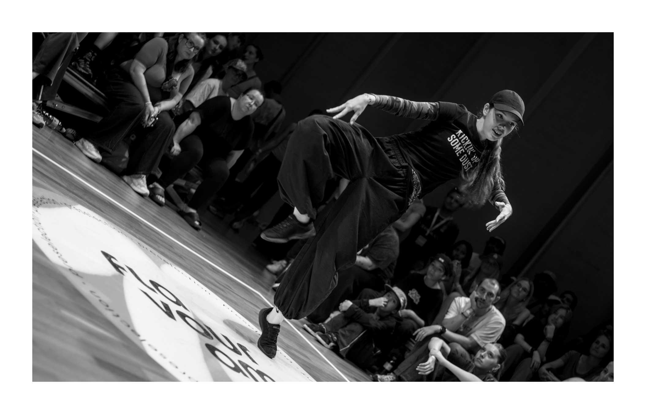 A black and white photograph of a dancer during a dance battle. She is standing on the point of her left foot, right raised and bent at the knee. The arms are spread with hands pointing down. It is akin a yoga or a martial pose. She is dressed in black from the cap all the way to the shoes. The far wall is in shades of blue and purple while the audience is out of focus, looking with rapt attention