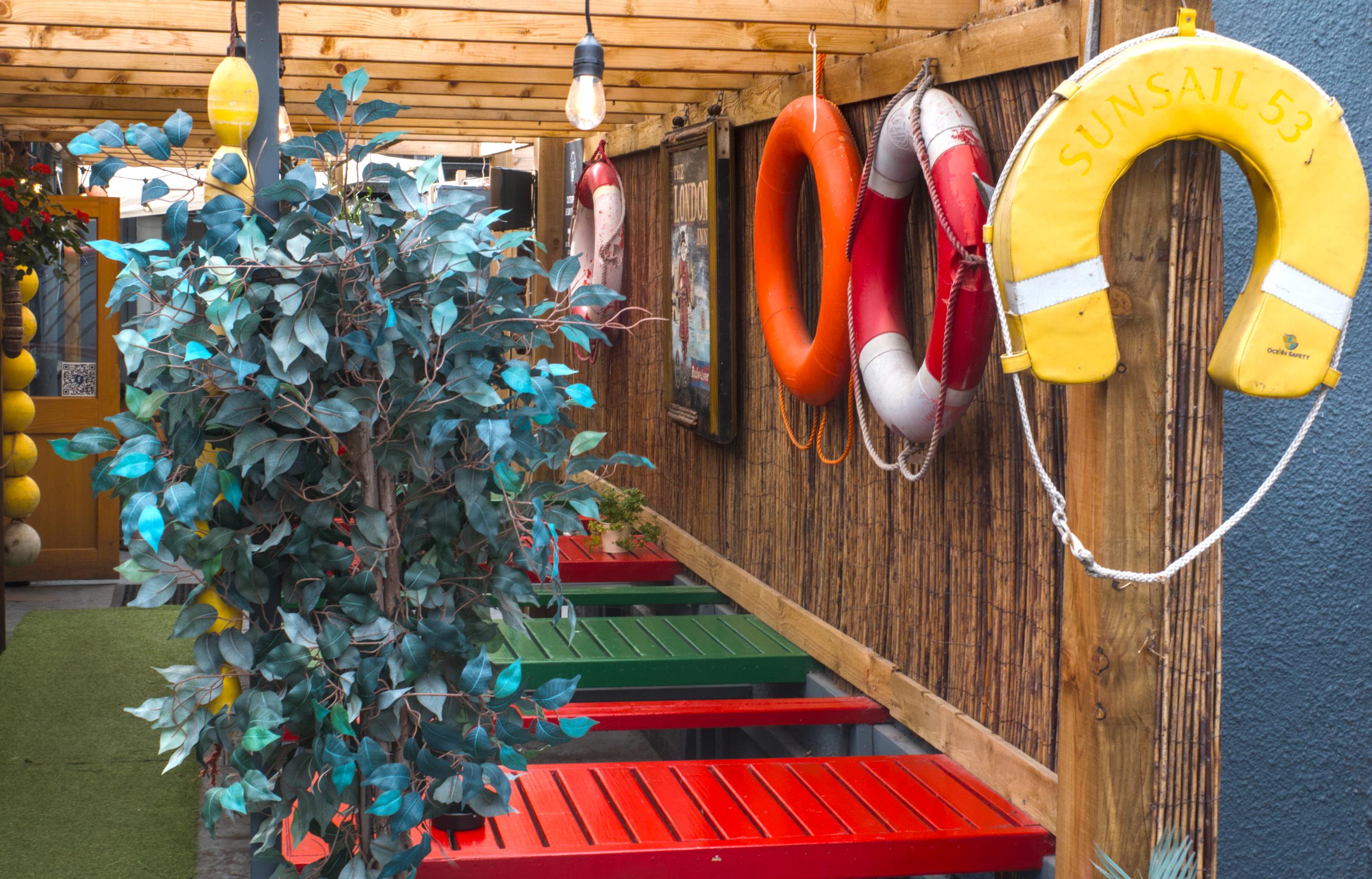 A colour photograph taken at the Captain's Cabin, Anchorhead, Weston Bay Somerset, UK
As I was passing by, I saw this open, colourful eating space and took this frame. Well it was so bright and cheerful it seemed rude no to.
Built of timbers, with a floor of artificial grass, red & green alternating tables & benches and decorated with different types of lifebelts.
