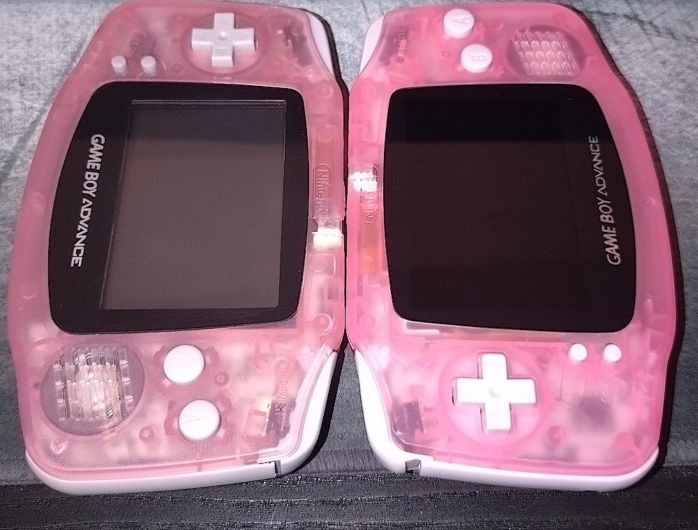 Two transparent pink Nintendo Gameboy Advance placed side by side on a surface. The consoles are positioned in such a way that they appear to be kissing each other.