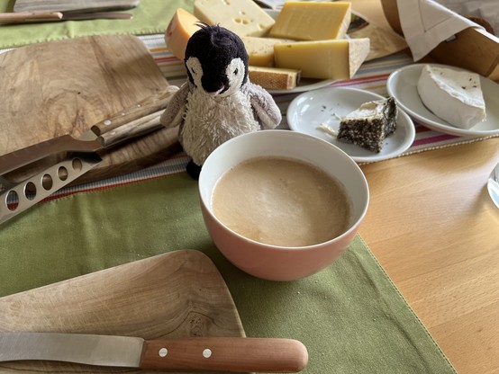 Ein gedeckter Frühstückstisch, zwischen Käse und einer Schale Milchkaffee sitzt eine Plüschpinguine