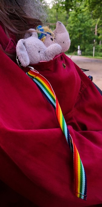 Die zwei kleinen Plüschies Einhorn Carola und Elefant Elfi in der Brusttasche eines bordeau-roten Hemdes, Elfis Regenbogen-Band hängt aus das Hemd herab.
