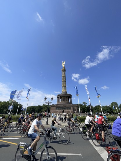 Eine große Gruppe von Radfahrern vor der Berliner Siegessäule an einem sonnigen Tag, mit blauen Flaggen und einem klaren Himmel im Hintergrund.