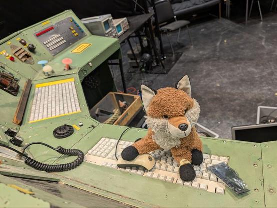 Ein Pluesch-Fuchs sitzt an einer nach Zeitmaschine ausschauenden Installation und bedient Maus und Tastatur. (und guckt dabei verschmitzt)