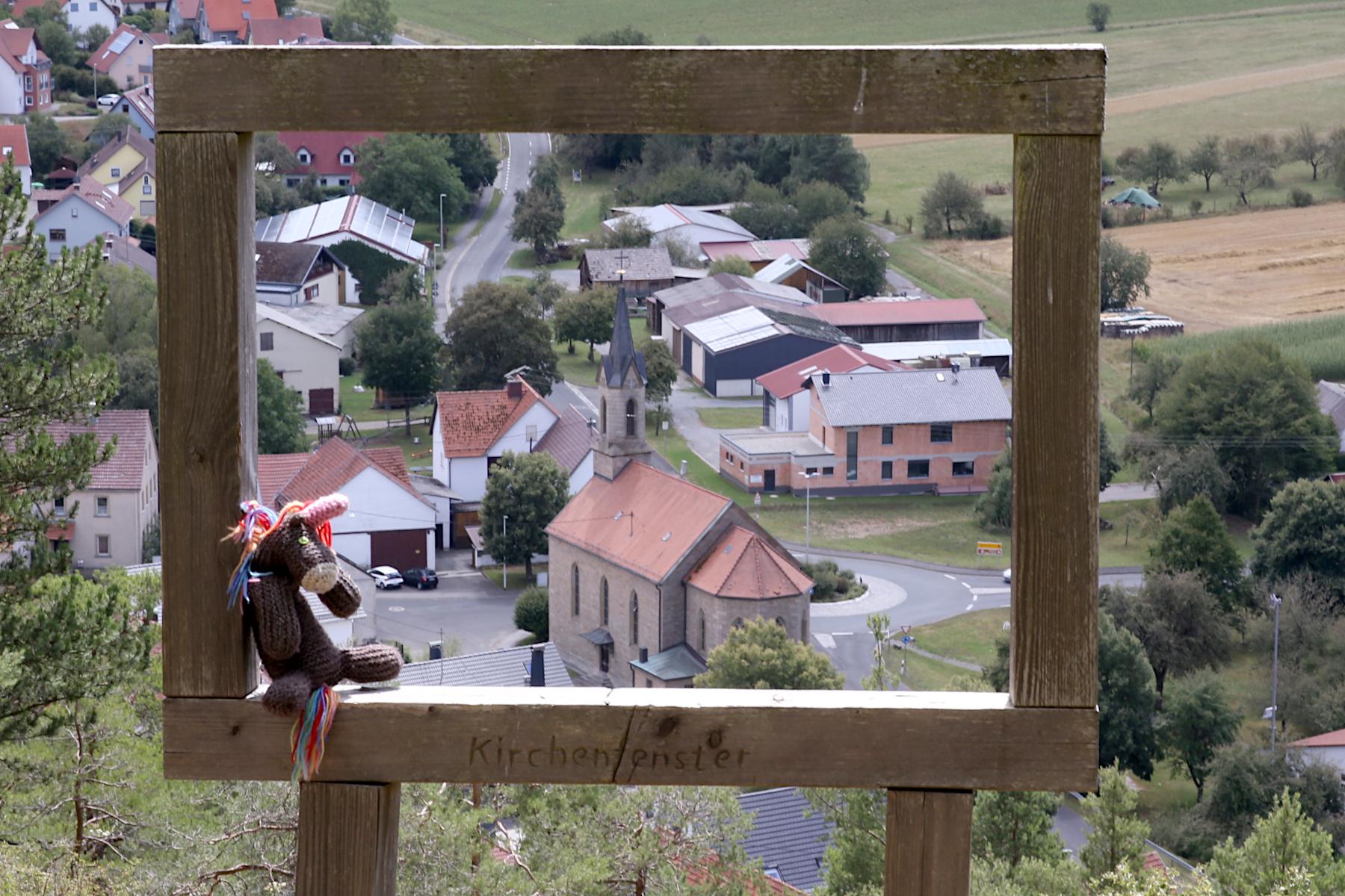 Max, das bunte Strickhorn, sitzt in einem hölzernen Bilderrahmen, beschriftet mit „Kirchenfenster“; im Hintergrund Blick auf die St. Michael-Kirche in Münnerstadt.
