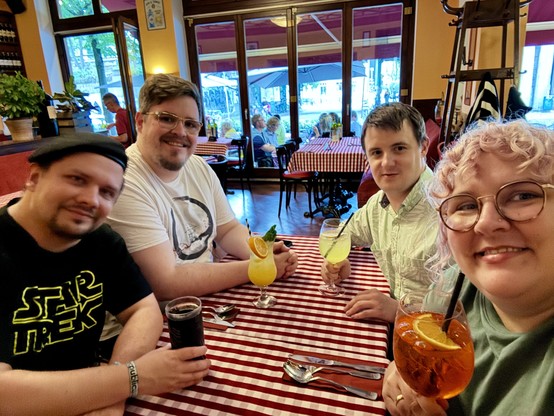 In einem italienischen Restaurant mit rot-karierten Tischdecken sitzen Macsnider, Christopher, Eric und Becci mit Spritz-Getränken um einen Tisch