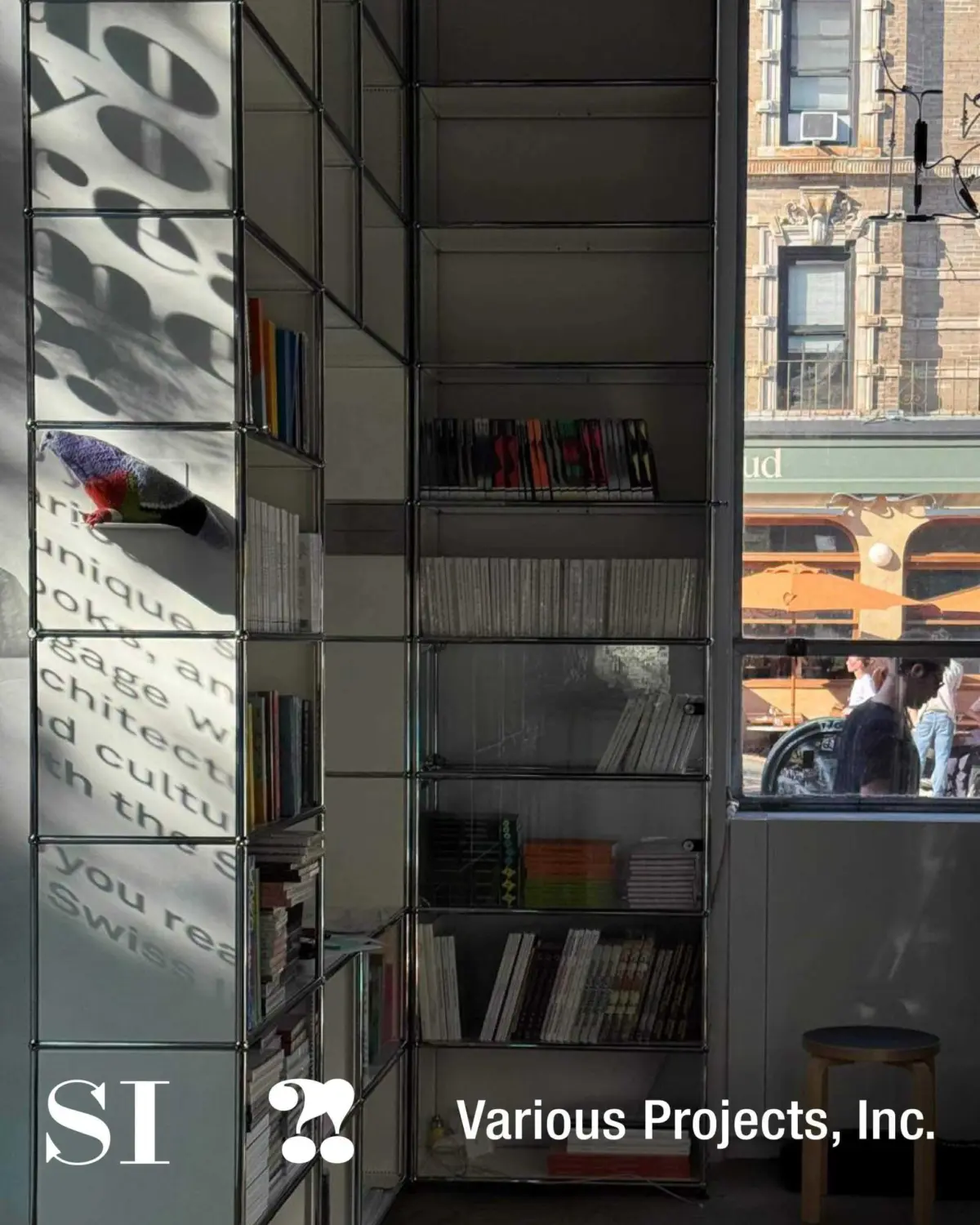 A modern, minimalist bookshelf display by Various Projects, Inc. The shelf features a mix of books, CDs, and vinyl records arranged in a sleek, industrial-style metal frame. The left side is occupied with shadows forming abstract text while the right side reflects a street scene with a storefront and pedestrians. The logo ‘SI’ and the text ‘Various Projects, Inc.’ are prominently displayed at the bottom left and right, respectively.