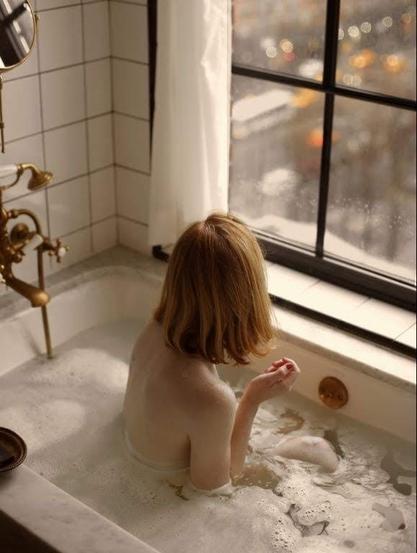 Girl see from behind in bath tub looking out of her window, staring at the traffic below her on the street.