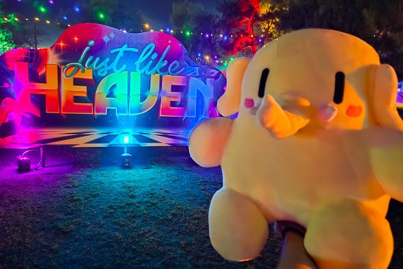 Photo of the Creature Mastodon stuffed toy in front of a ”Just Like Heaven” festival sign during the night of the festival. The Creature is facing the camera in the foreground towards image-right. In the background is the human-height sign lit up in neon magenta, cyan, and purple lights installed on the surrounding grass lawn. Further in the background are palm trees and more festival lights.