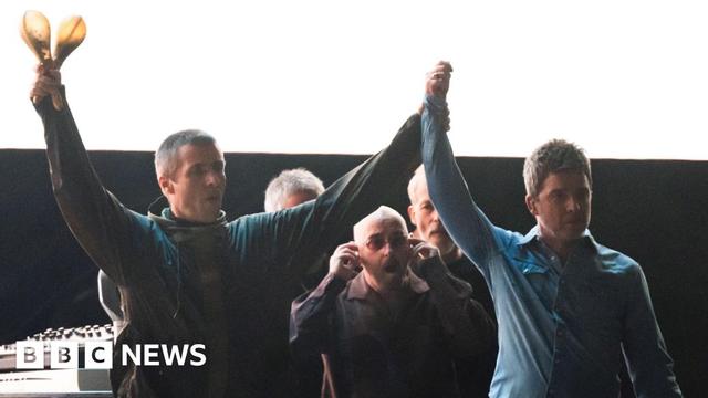 Liam and Noel hold their hands up in the air on stage