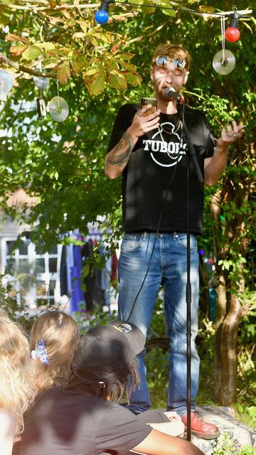 William Sommer læser op fra sin smartphone på en scene iført Tuborg t-shirt, jeans og solbriller. Det er sommer under et træ, hvor der hænger farvede lys og cd'er