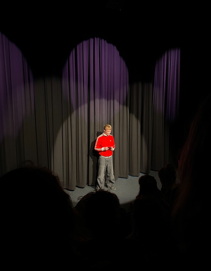 Christian Enevoldsen står på en teater-scene i spotlyset, iført skrigrød Adidas trøje og læser op.