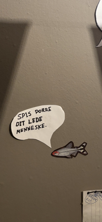 Billedet viser en væg med en udklippet tegning af en fisk og en taleboble, som er klistret op. Taleboblen indeholder teksten:  “SPIS PORRE DIT LEDE MENNESKE.”