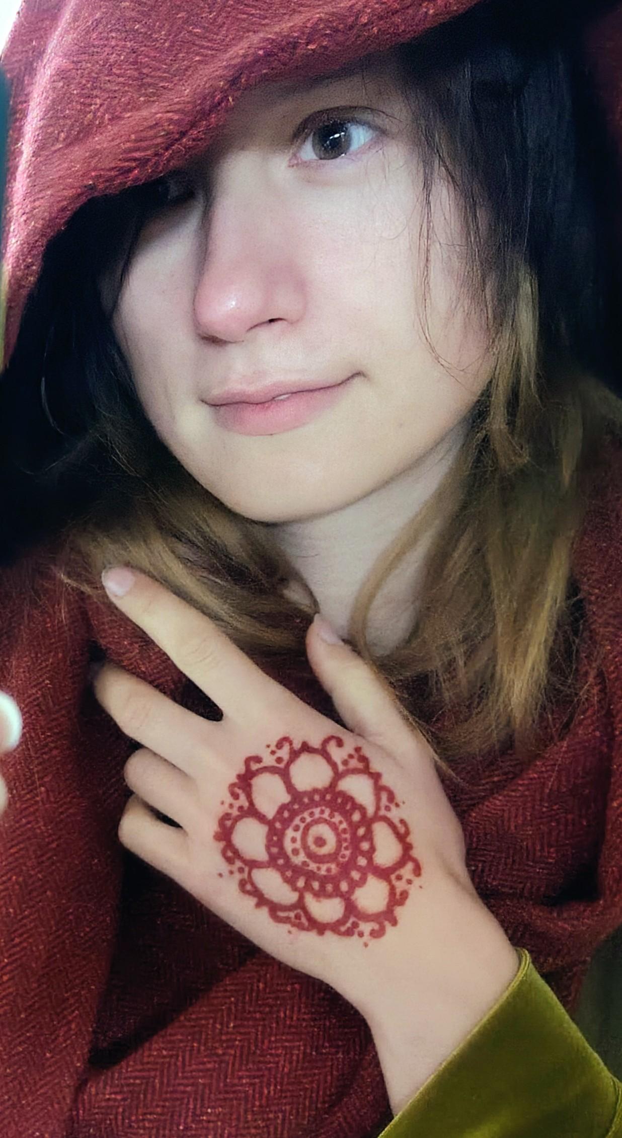 Person (me) with a red wool hood scarf on, holding their hand over their collar, showing red henna patterns resembling a passionflower on their hand