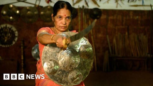 A woman wearing an orange saree stands with a brass shield and a sword