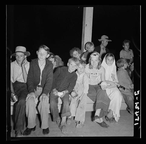 The image is a black and white photograph showing a group of people sitting on benches in an outdoor setting at night. There are both adults and children, with most wearing casual or work-related attire such as overalls, hats, jackets, and shoes typical for the era suggested by their styles. Some individuals appear to be engaged in conversation while others look off into space or directly towards the camera.

The people have a range of expressions; some seem contemplative or attentive, while others exhibit more relaxed demeanors with smiles on their faces. The lighting is low, likely from artificial sources given its nighttime setting, which casts shadows and creates contrast among the individuals in the group.

In the background, there's an open space leading to what appears to be a larger area where additional people are standing or seated under other light sources not visible within this frame. A wooden post stands out prominently on one side of the image, likely part of a structure such as a fence or barrier that surrounds them. The overall atmosphere suggests a communal gathering at an event like Halloween mentioned in the provided context.

The photograph is dated "13-4/25," which may refer to April 1948 (as inferred from the format typically used for photos and documents during this time). This specific image captures a moment of social interaction among individuals who might be part of a migratory or community gr [...]