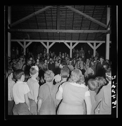 The black and white photograph captures a lively scene of people gathered in what appears to be an open-air pavilion or barn during the evening. The crowd consists mainly of adults, with some children visible towards the front. Many attendees are dressed casually; men wear hats while women sport headscarves or hair accessories. Some individuals have on costumes that suggest a festive atmosphere.

The focus is not clear enough to discern specific details about facial expressions and interactions among participants due to motion blur and camera shake typical of night photography from mid-20th century America. The image's monochromatic palette emphasizes the social gathering over individual identities, capturing the communal spirit rather than personal characteristics.

In terms of context, this photo is attributed to Dorothea Lange who documented various aspects of American life during her career as a photographer for both the Migrant Farm Workers and the Farm Security Administration. The caption "Halloween party at Shafter migrant camp" indicates that this image likely depicts an event celebrating Halloween with migrants in California.

The visible text on the right side, possibly part of the photograph's border or watermark, reads 'Lange,' perhaps a signature or label by Lange herself or from her collection, adding authenticity to the historical context.
